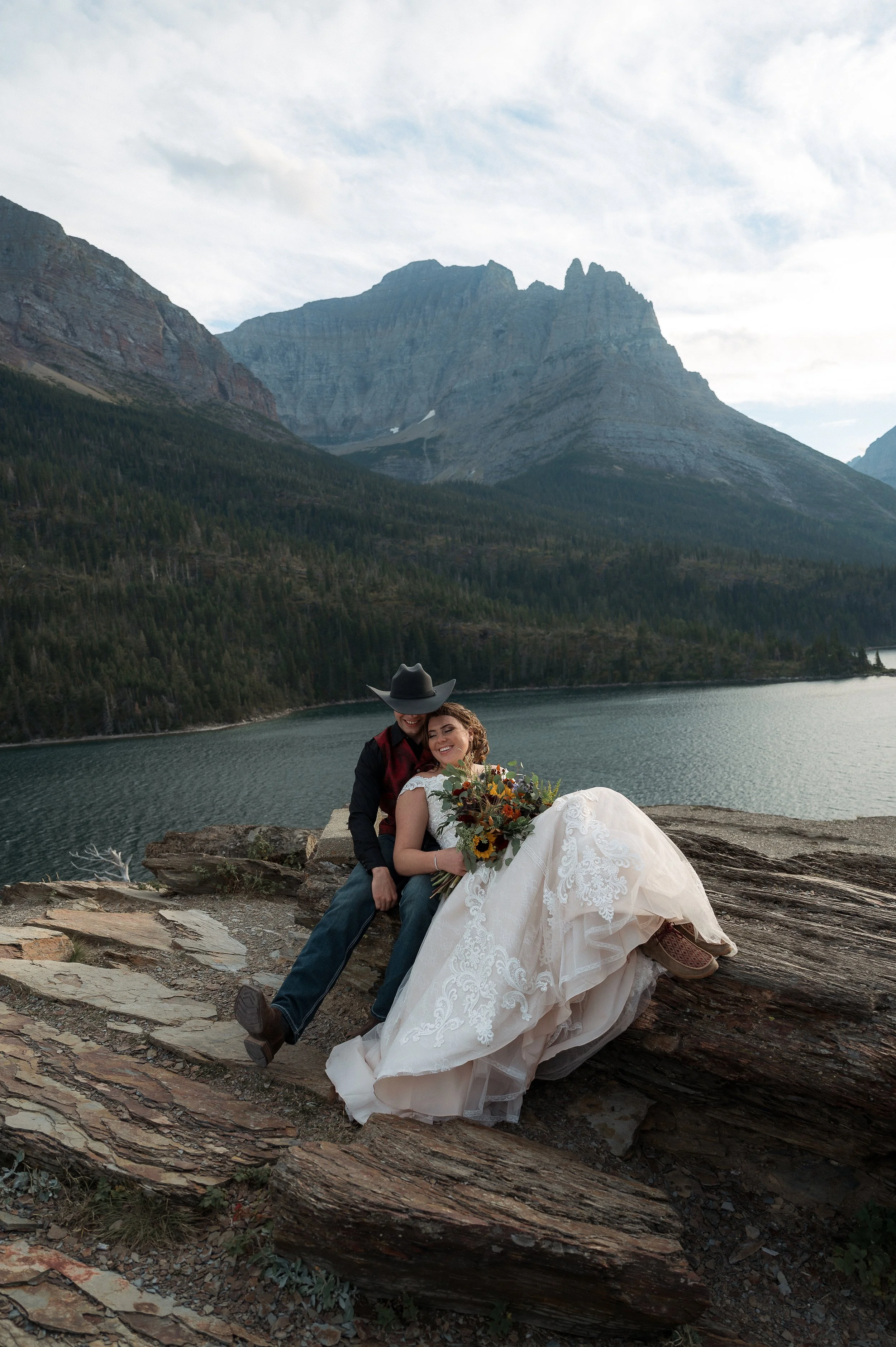 A wedding couple sitting on rocks near a lake, with mountains and a cloudy sky in the background. The bride is in a white dress holding a bouquet, and the groom is wearing a hat and casual attire.