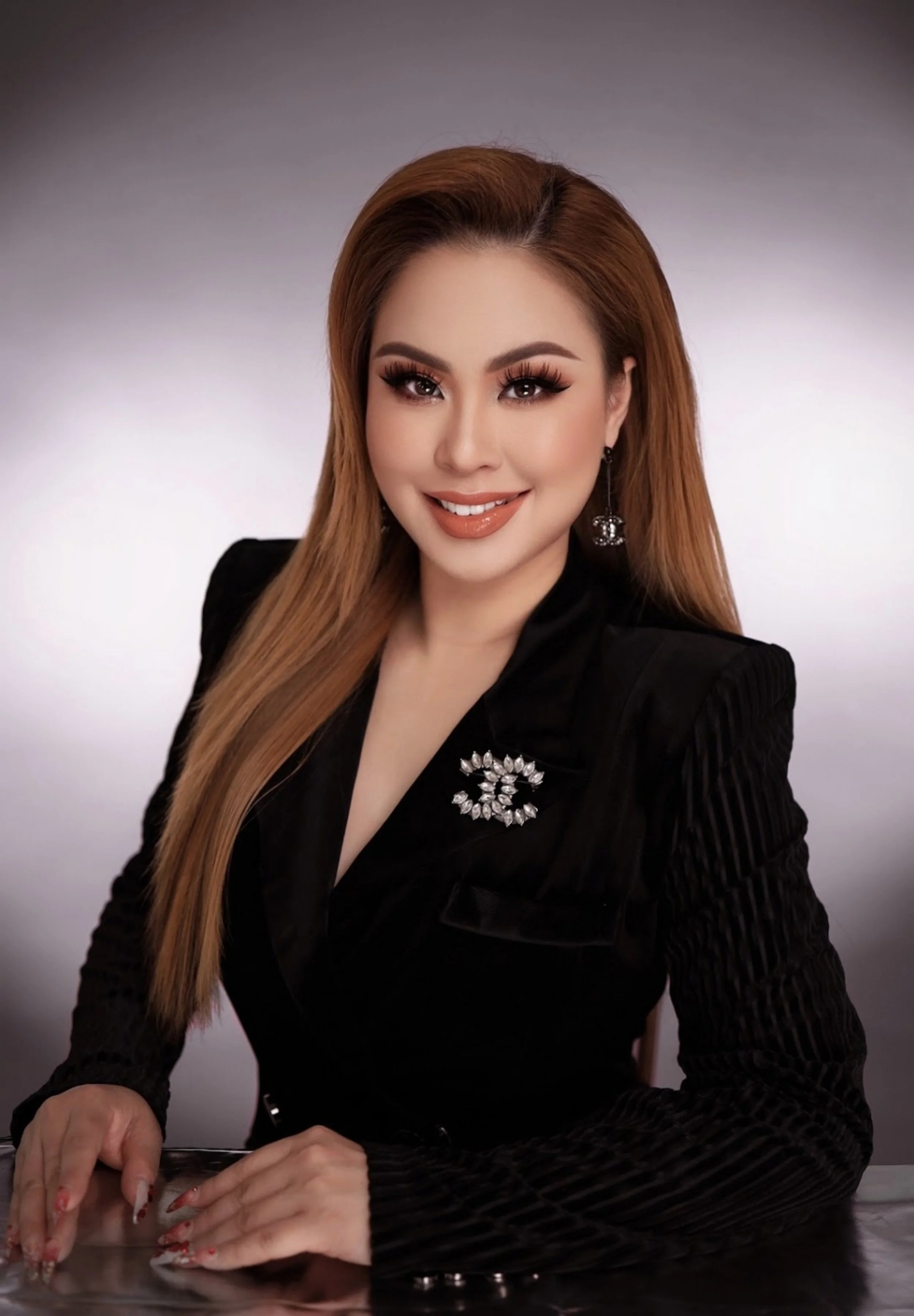 A woman with long light brown hair, wearing a black blazer with a rhinestone brooch and earrings, smiling at the camera against a gray background.