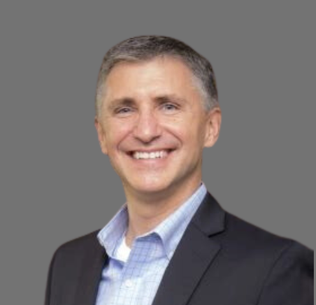 A smiling man in a suit with a light blue shirt against a gray background.