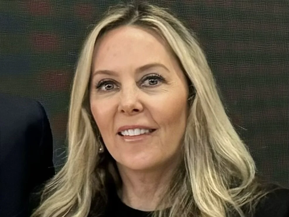 A woman with blonde hair smiling, wearing makeup and earrings, in a dark-colored top, with a blurred background.