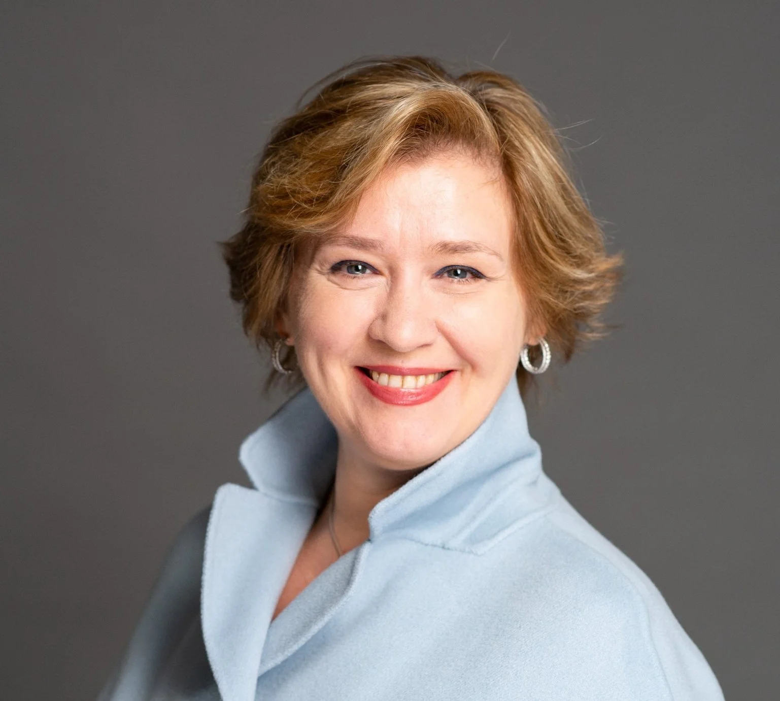 A woman with short, wavy, light brown hair smiling, wearing a light blue high-collared coat and silver hoop earrings, posed against a plain gray background.