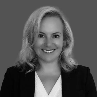 A woman with shoulder-length blonde hair and a friendly smile, wearing a black blazer and a white top, posing against a plain gray background.