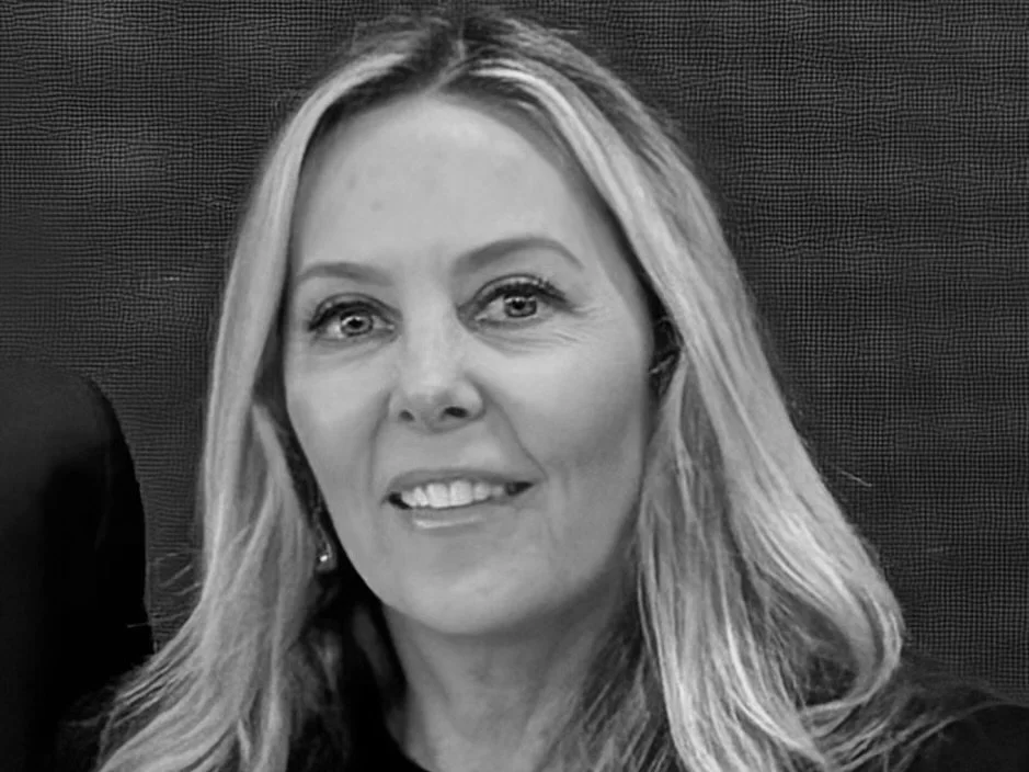 Black and white photo of a woman with long blonde hair, smiling with her teeth showing, against a dark background.