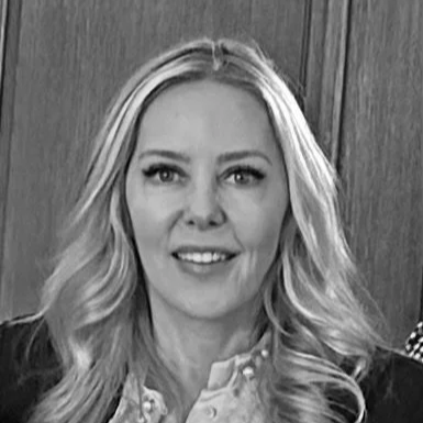 A woman with long blonde hair wearing a black blazer and light pink blouse, smiling against a gray background.