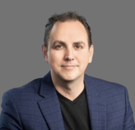 A man with short dark hair and light skin wearing a dark blue blazer and black shirt, smiling against a gray background.