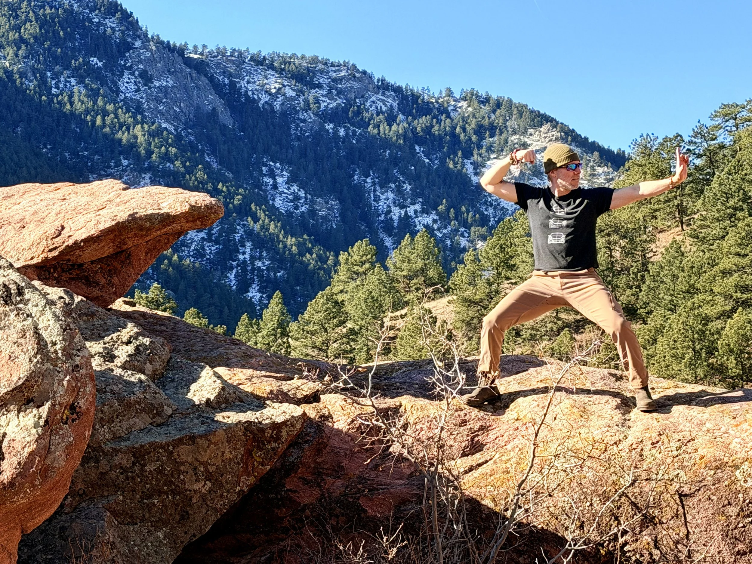 Eran reznik preforming movement from the Ba Duan Jin Qigong form - Stretch the bow to shoot the eagle , standing on a boulder with a view of snowy mountains and pine trees in the background
