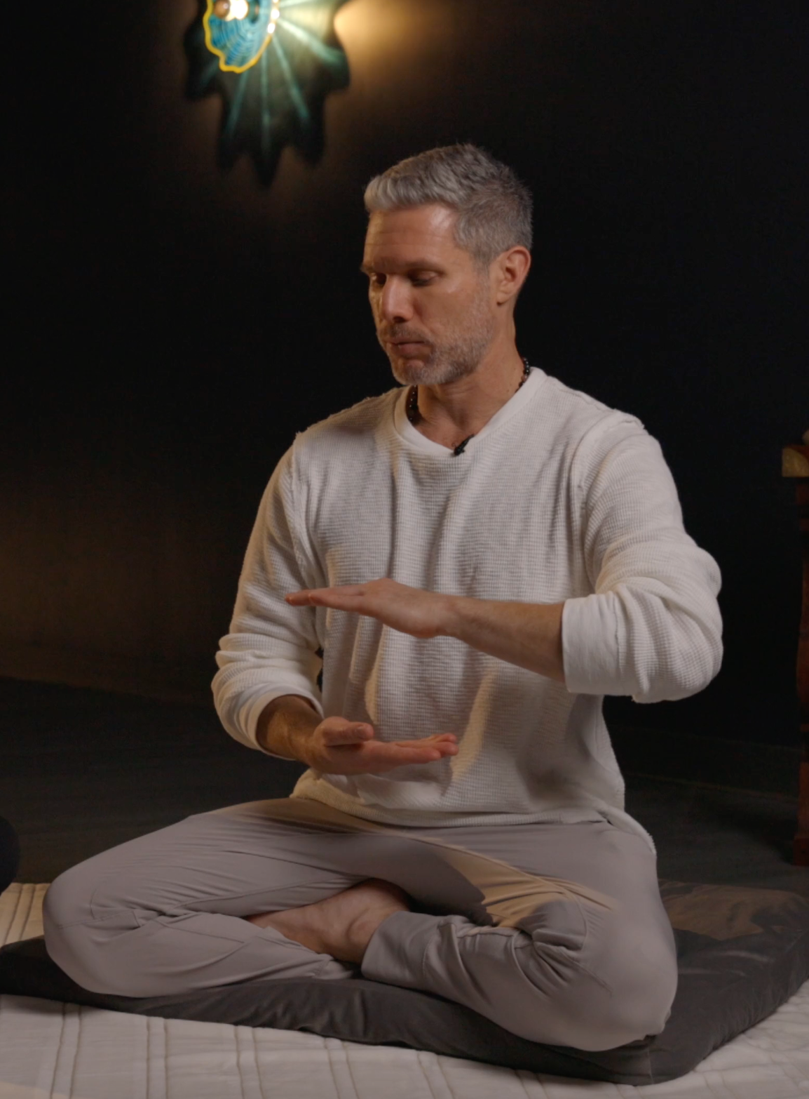 A middle-aged man with gray hair and a beard, sitting cross-legged on a cushion, practicing meditation or mindfulness with his hands positioned as if holding an object. He is wearing a long-sleeve beige shirt and beige pants, in a dimly lit room with a dark wall behind him, in a serene or spiritual setting.