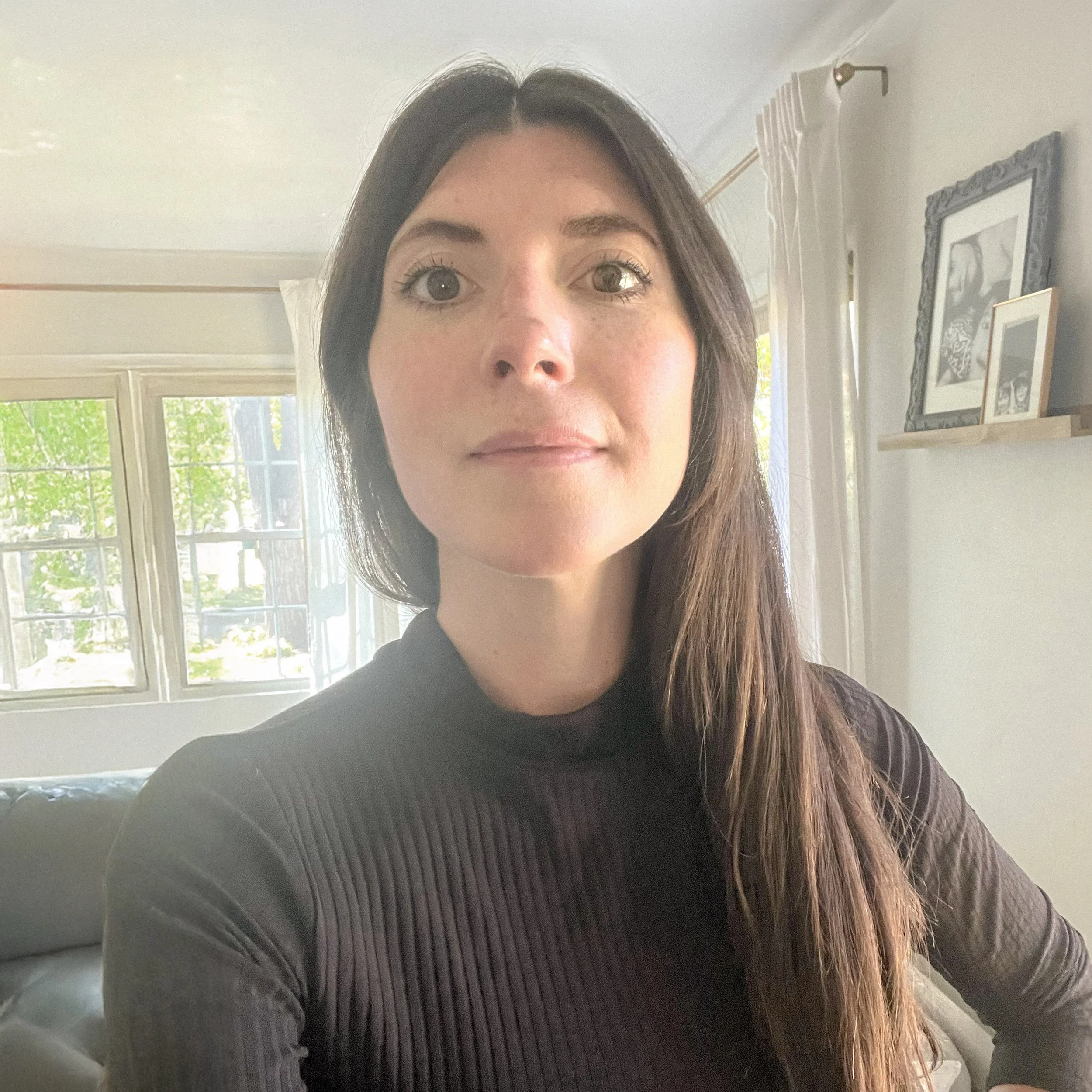 A woman with long brown hair taking a selfie in a well-lit room with large windows and framed pictures on the wall.