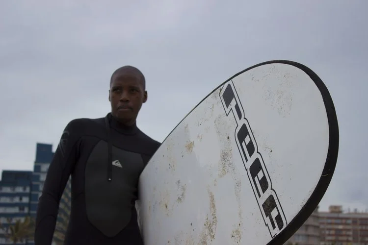 Man in black wetsuit holding a white surfboard with dirt marks, standing outdoors on a cloudy day, with buildings in the background.