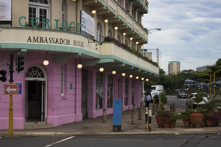 Pink and beige building labeled 'Ambassador House' with decorative lighting, windows, plants, and sidewalk street scene