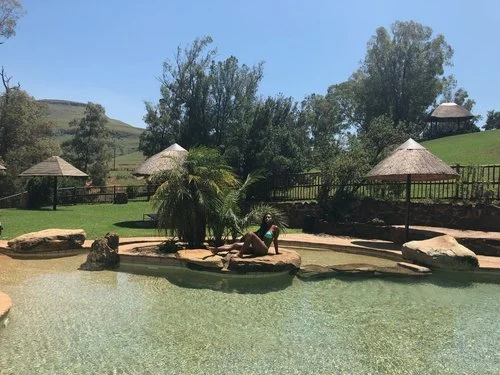 Person lounging on rocks near a pond at a park with trees, grass, and gazebo structures in the background on a sunny day.