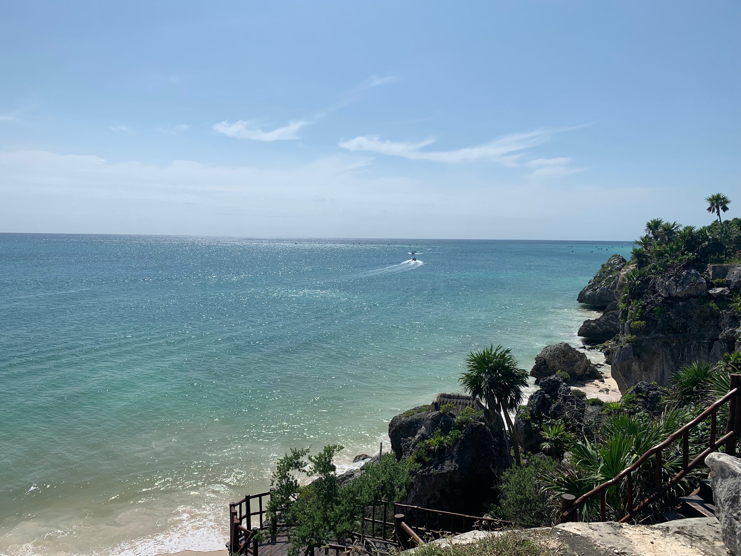 A tropical beach with clear turquoise water, rocky coastline with palm trees, and a boat speeding across the ocean under a partly cloudy sky.