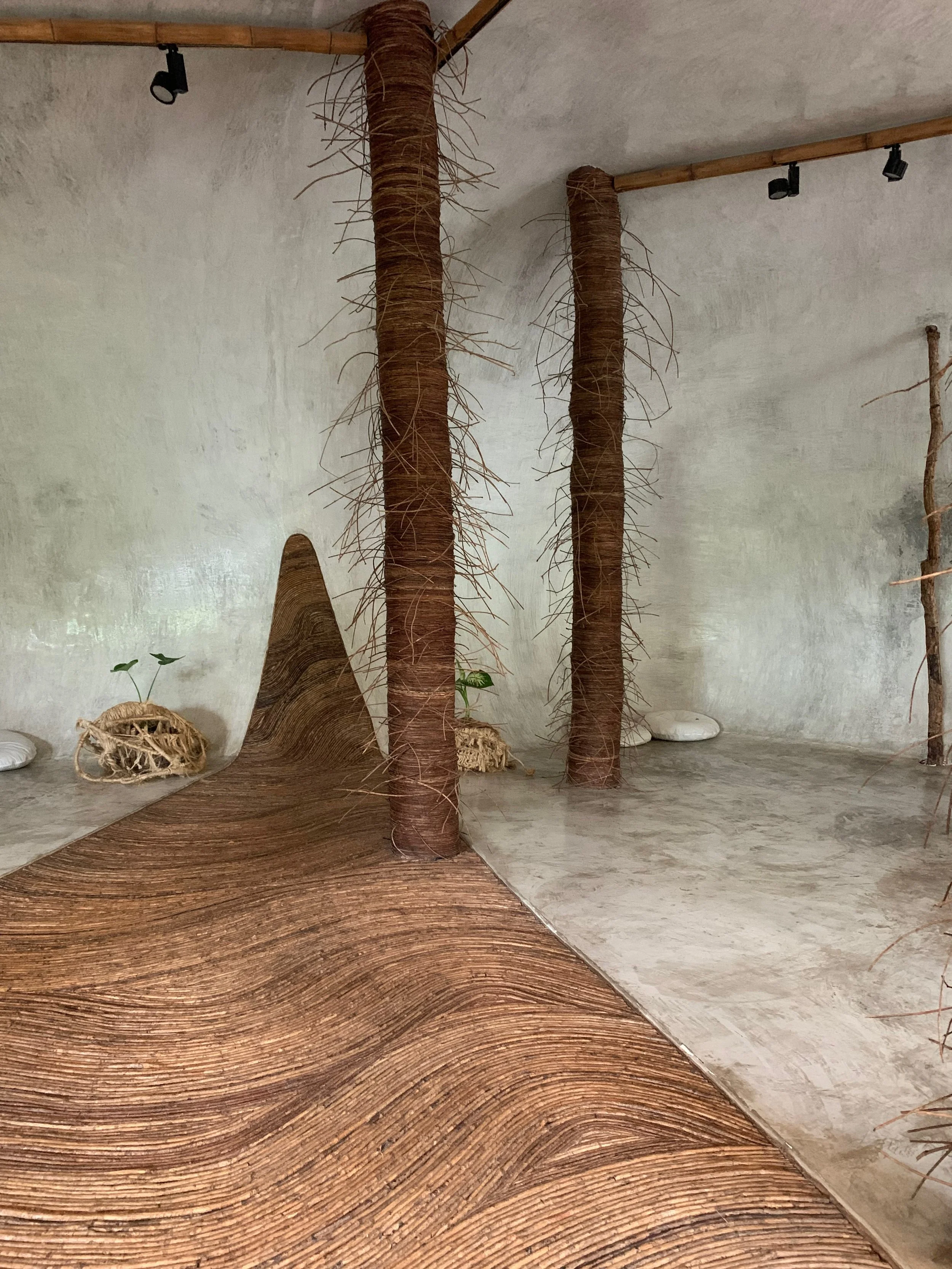 Interior space with two large tree-like structures made of woven material, a small plant in a sack on the floor, and a curved wooden surface with a textured pattern, against plain concrete walls.