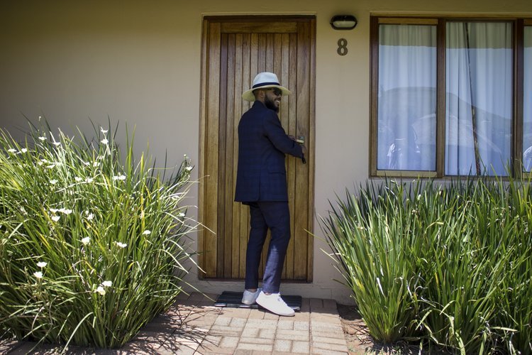 A man wearing a white hat, sunglasses, navy blazer, navy pants, and white sneakers is standing at a wooden front door of a house, holding the door handle and smiling.