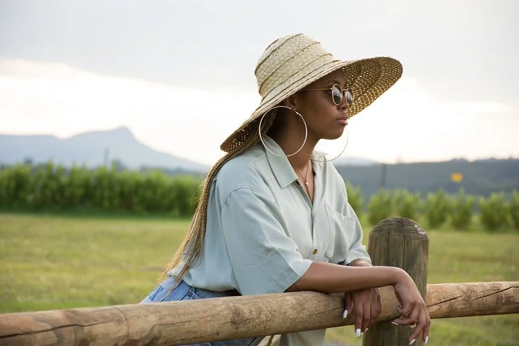 Woman with sunglasses and a wide-brimmed straw hat leaning on a wooden fence, looking into the distance, with greenery and mountains in the background