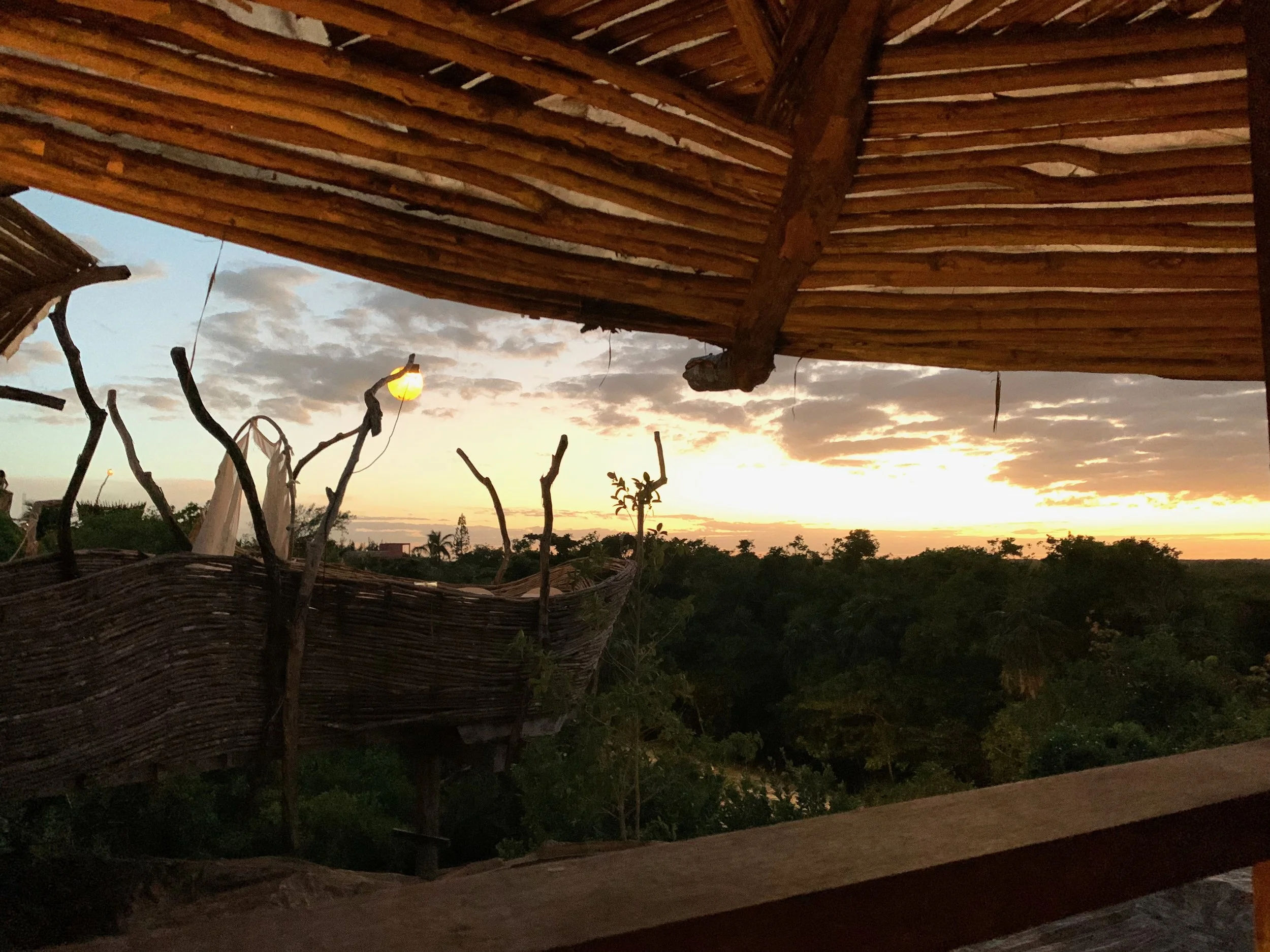 A rustic wooden balcony overlooking a lush landscape at sunset with a partly cloudy sky.