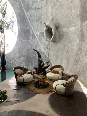 Indoor seating area with four round cushioned chairs arranged around a small round table on a woven rug, with a large decorative wall sculpture and an artificial plant in the background.