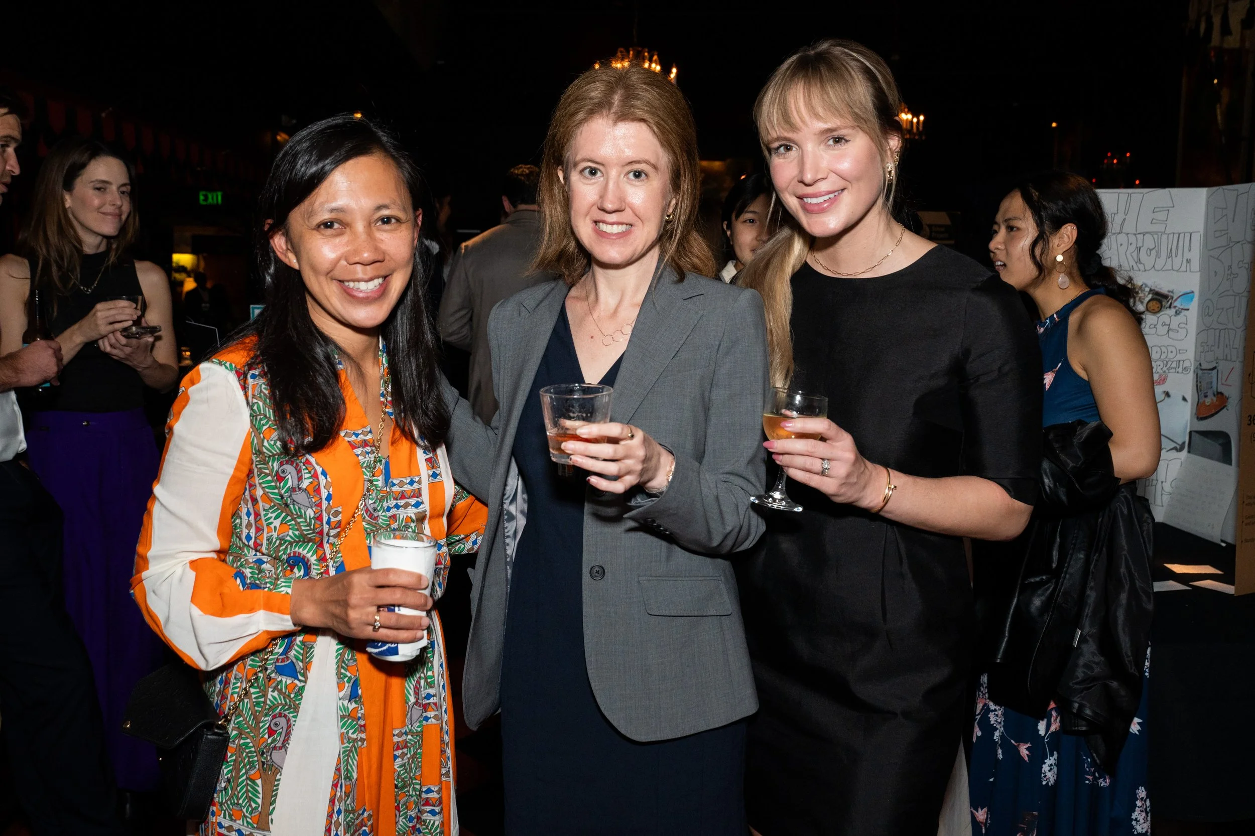 Group of women at a social event, smiling, holding drinks, in a dimly lit indoor venue.