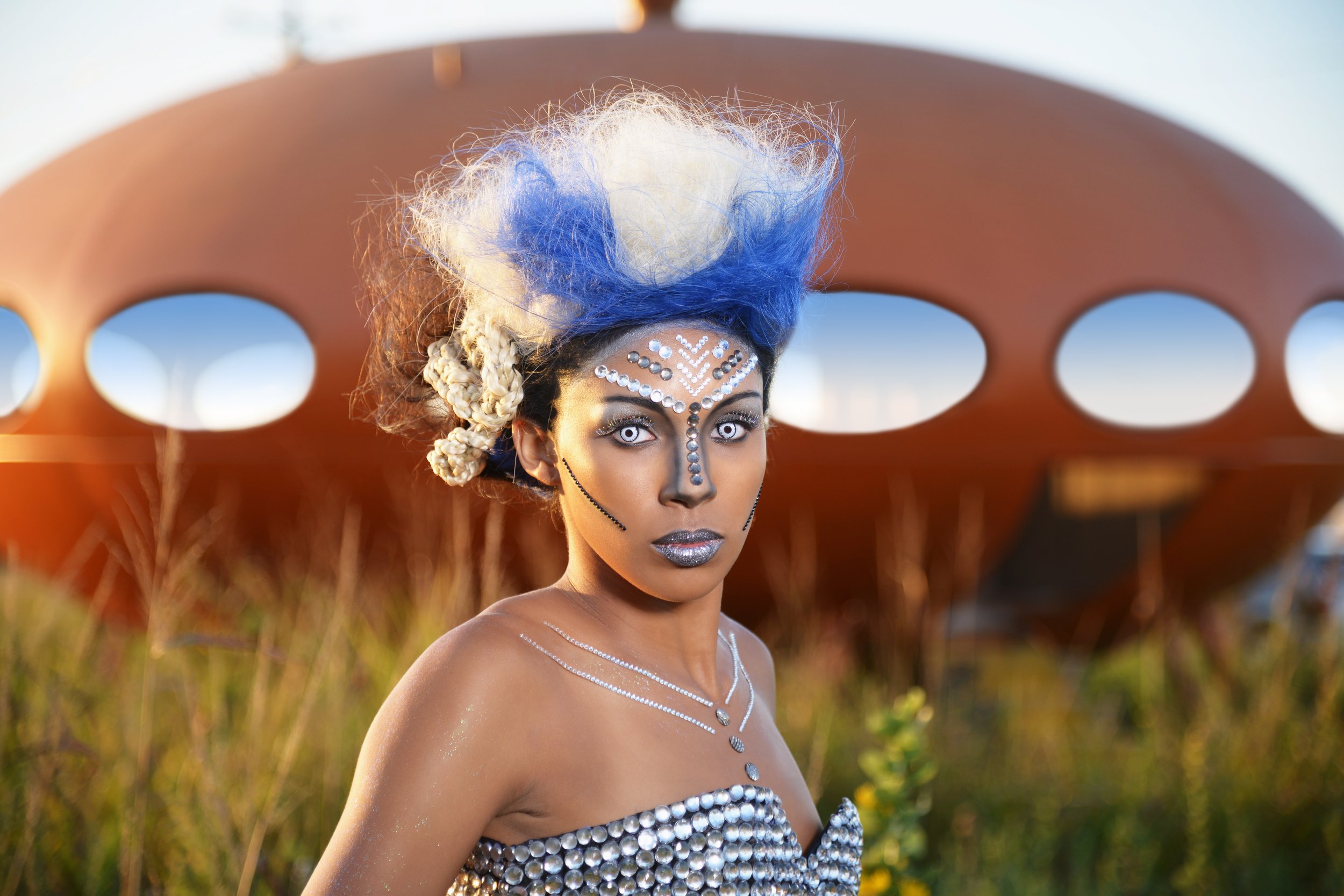 A person with blue and white hair and elaborate silver makeup poses outdoors in front of a futuristic, dome-shaped building with circular windows. They are wearing a metallic studded outfit.