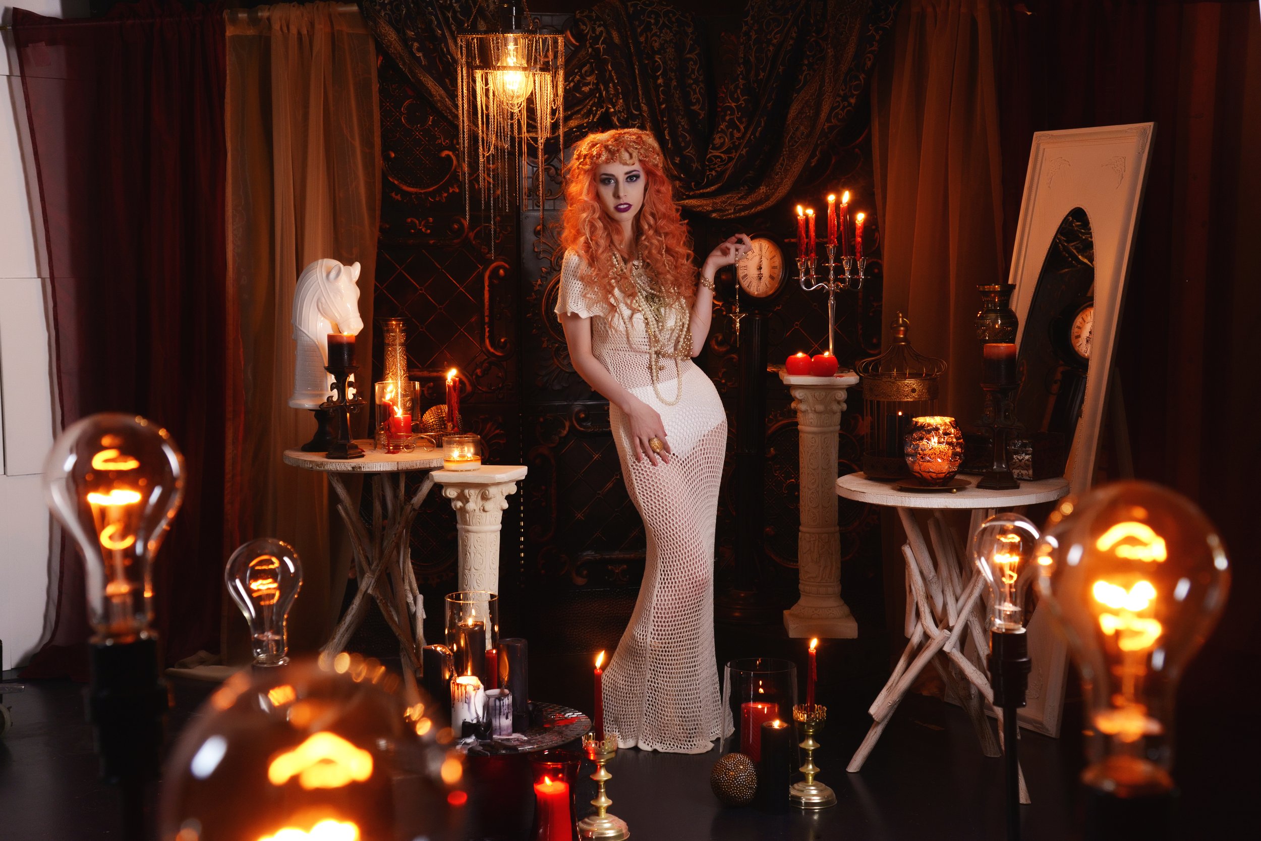 A woman with curly red hair stands in a dimly lit, decorated room with vintage decor. She wears a long, white, see-through dress. The room is adorned with lit candles, an ornate clock, a white horse bust, and a draped mirror. The lighting includes ha