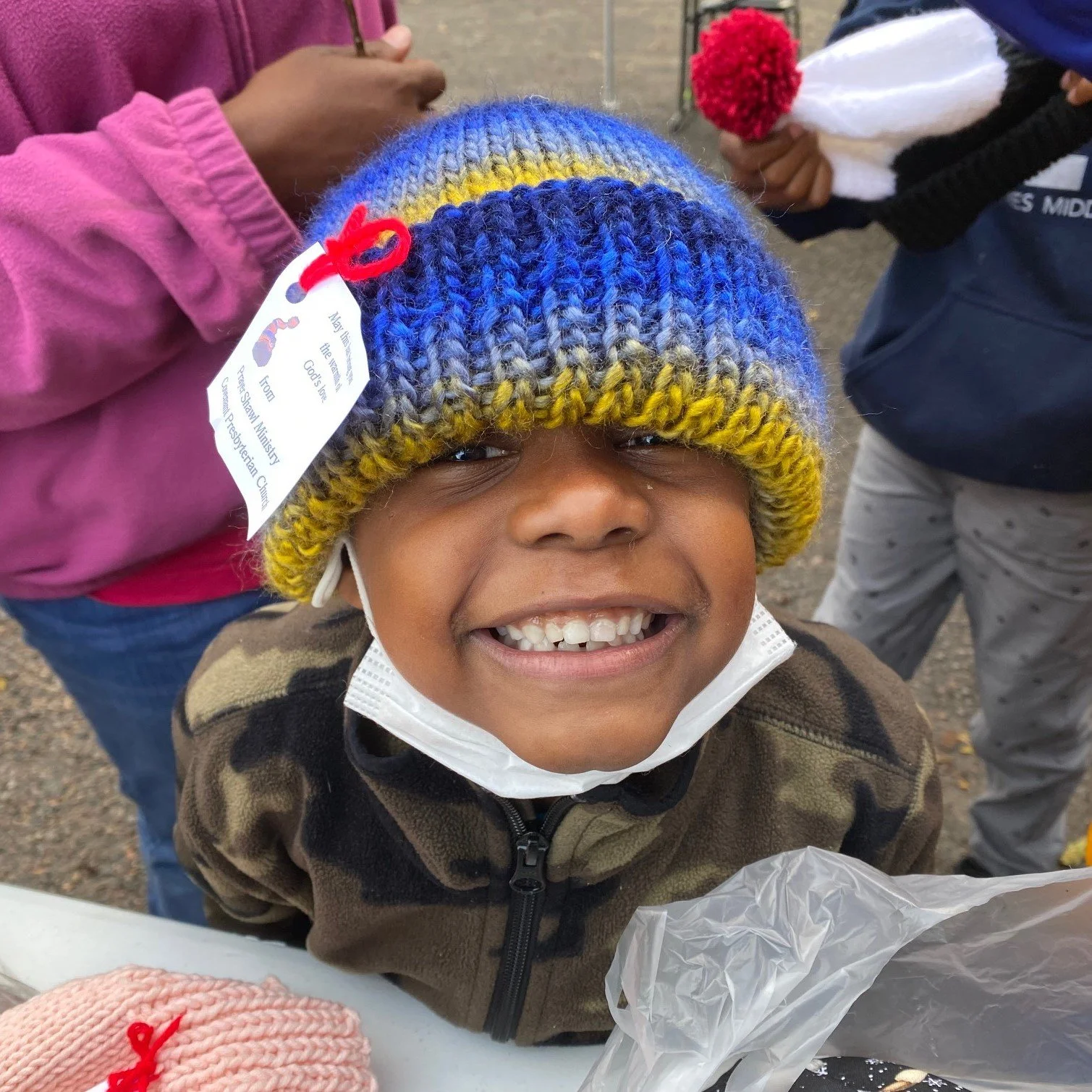 A smiling child wears a blue, gray, and yellow knitted hat.