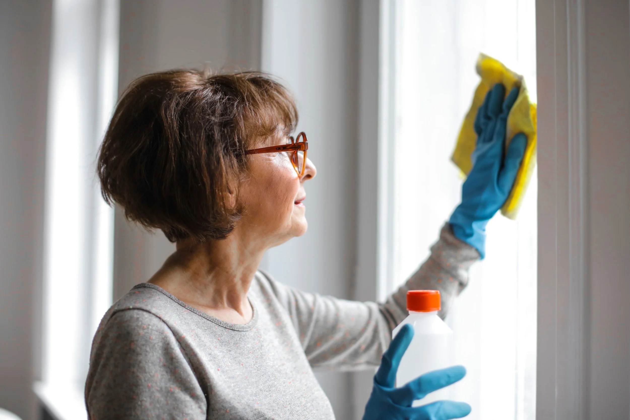 Frau mit Brille putzt ein Fenster und trägt dabei blaue Gummihandschuhe.