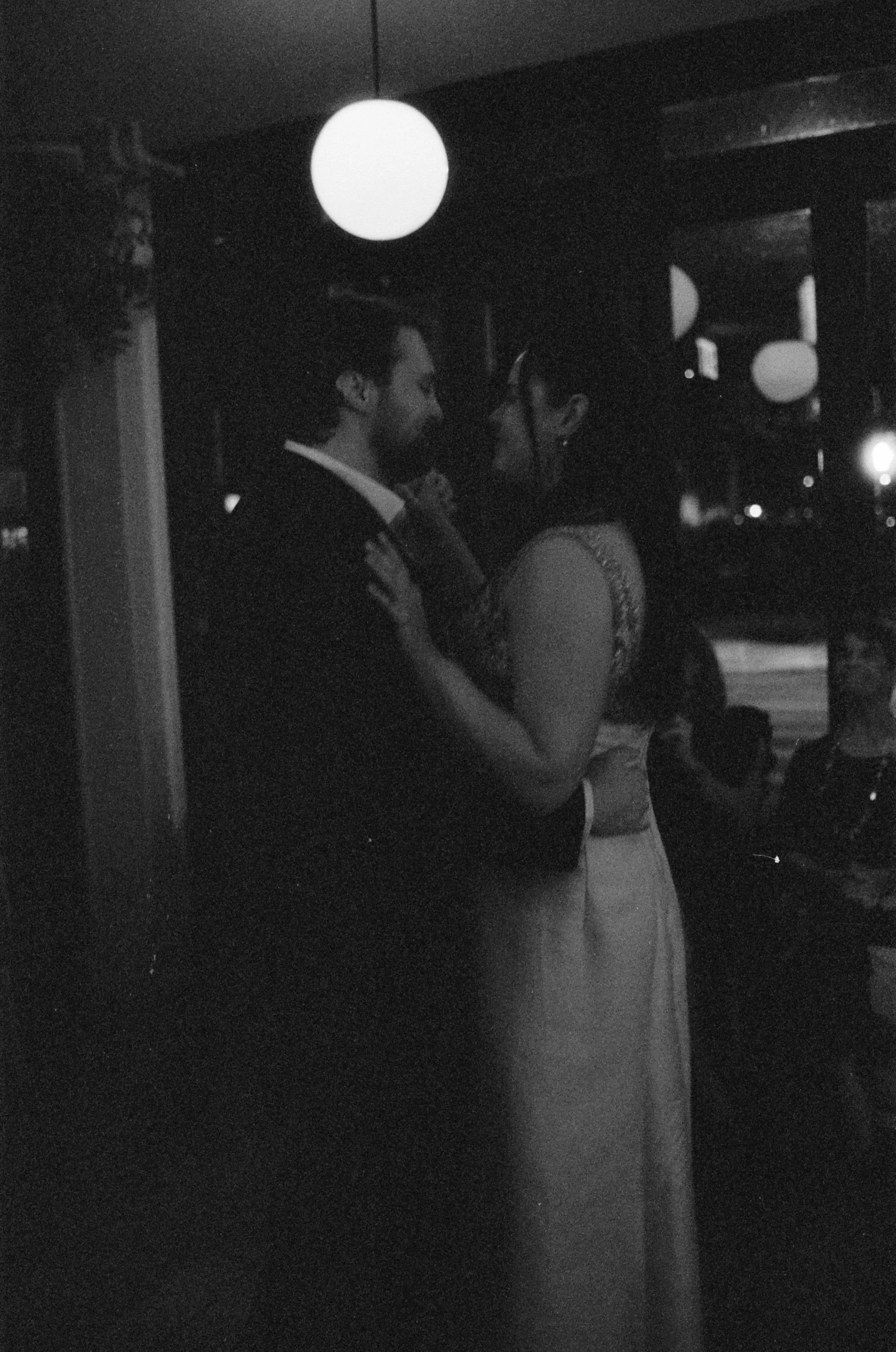A couple dancing closely in a dimly lit setting, with round ceiling lights and other people seated in the background.