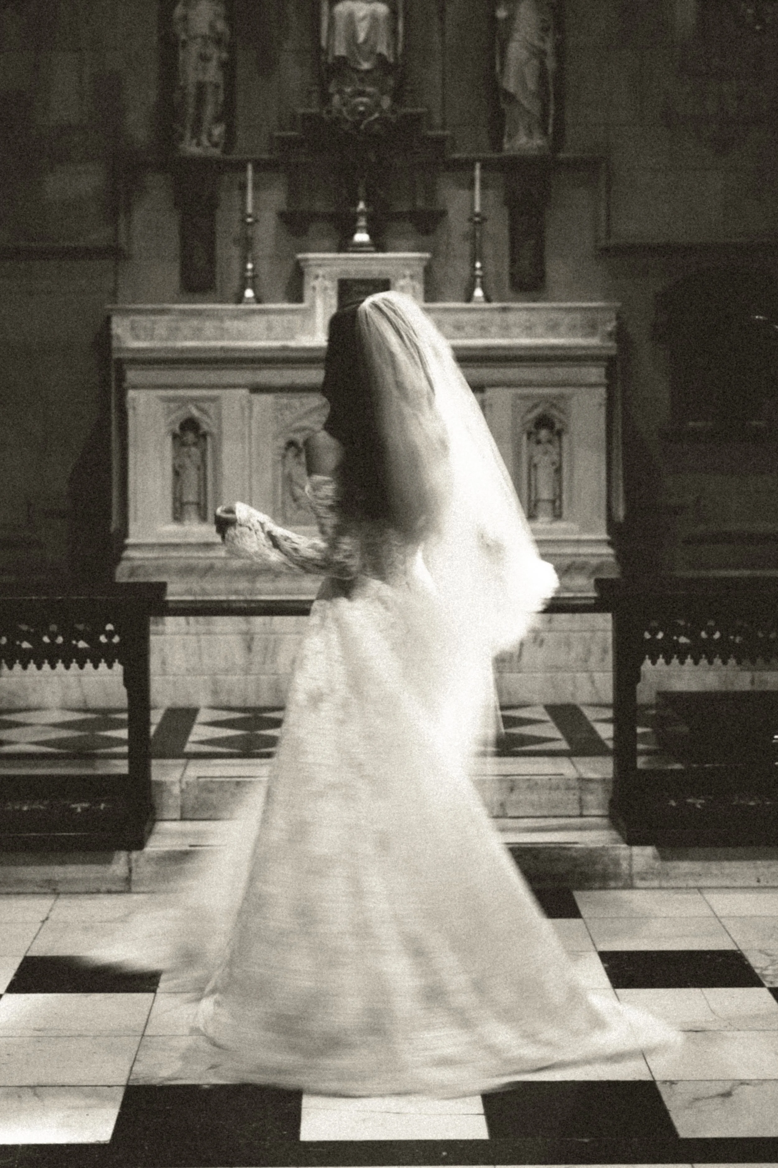 A woman in a wedding dress with long hair is standing inside a church, facing the altar, holding a small bouquet of flowers. The church has an ornate altar with candles and religious artwork.