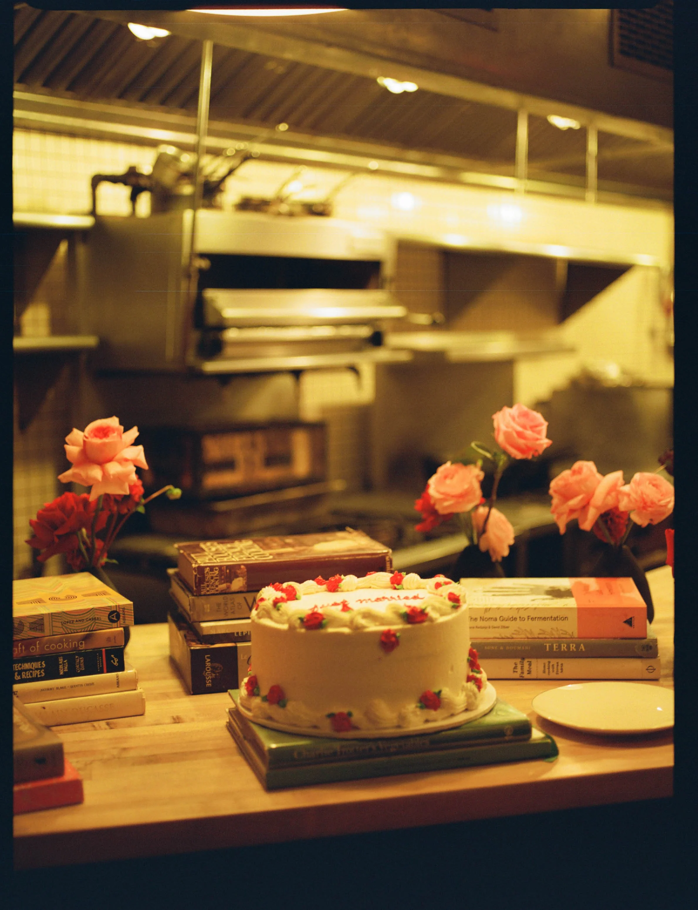 A birthday cake with strawberries and cream on a wooden table, surrounded by books and pink flowers, in a kitchen setting.