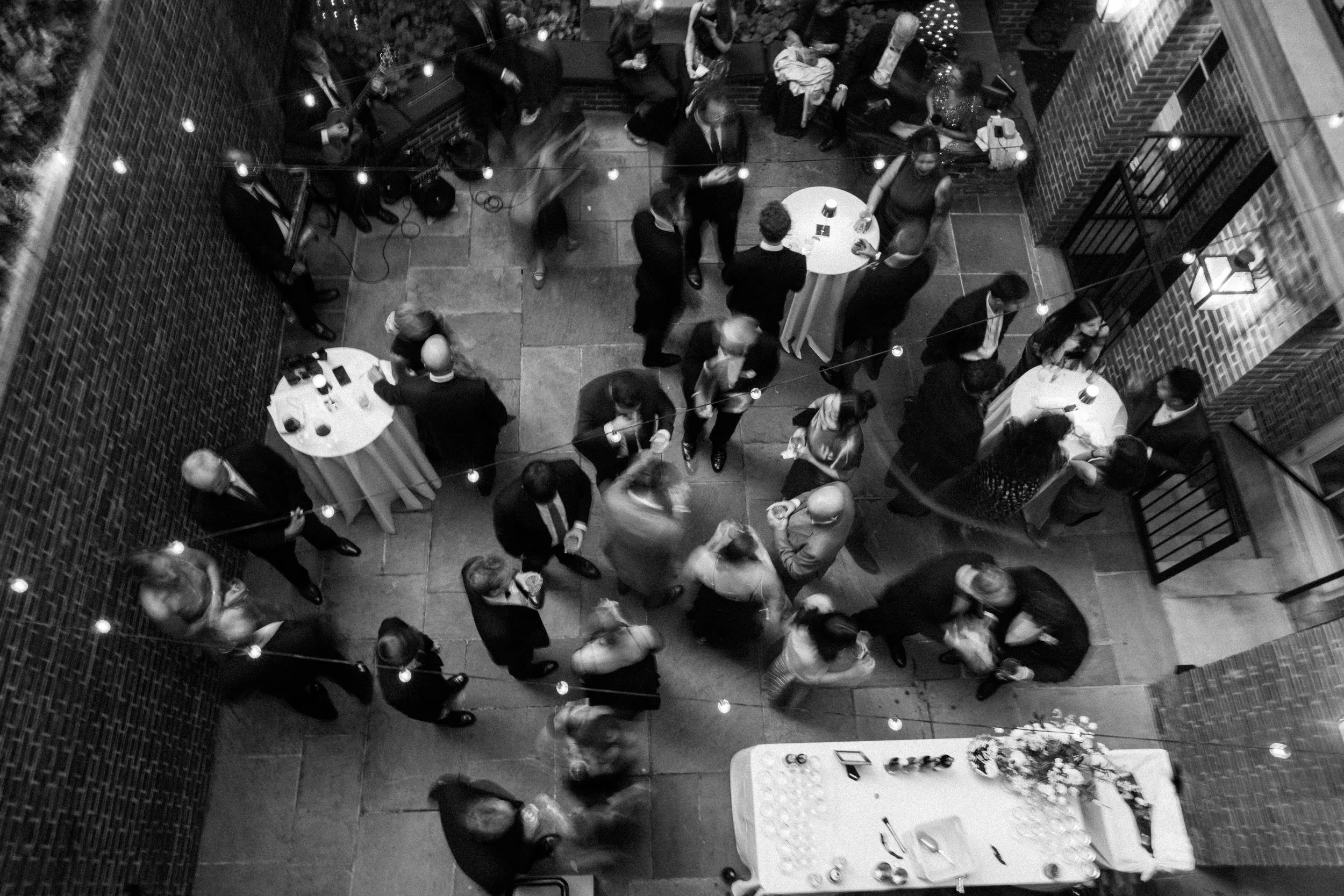 A black-and-white photo of a social gathering, showing people mingling, some standing around small round tables, others dancing, with strings of lights overhead and a musician playing guitar in the corner.