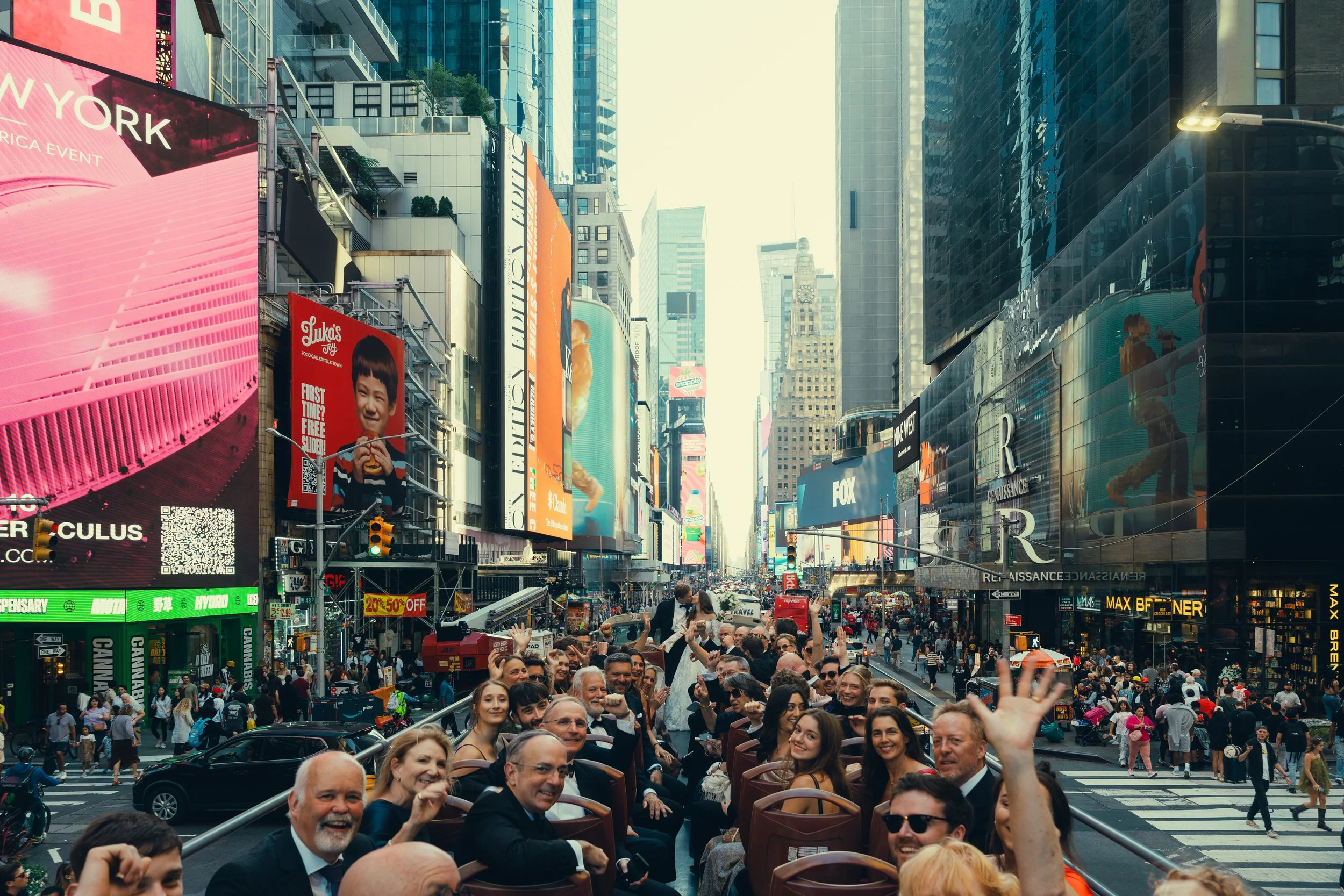 A lively scene in Times Square, New York City, with a large crowd of people, aerial tour bus with seated tourists, illuminated billboards, and tall skyscrapers.
