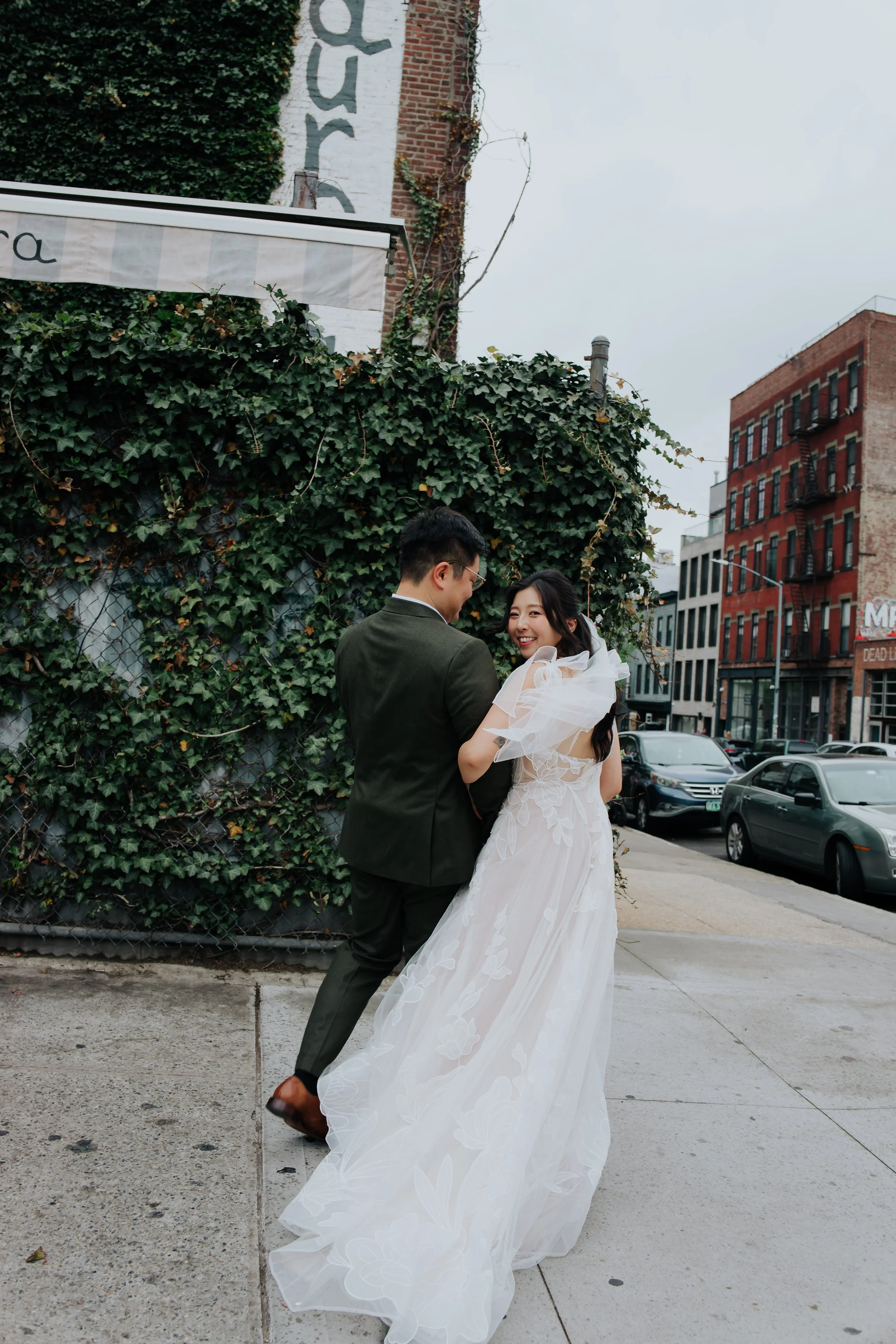 A bride and groom standing outdoors on a city sidewalk, smiling at each other. The bride is wearing a white wedding dress, and the groom is in a dark suit. Behind them is a brick and ivy-covered wall and several parked cars in the background.