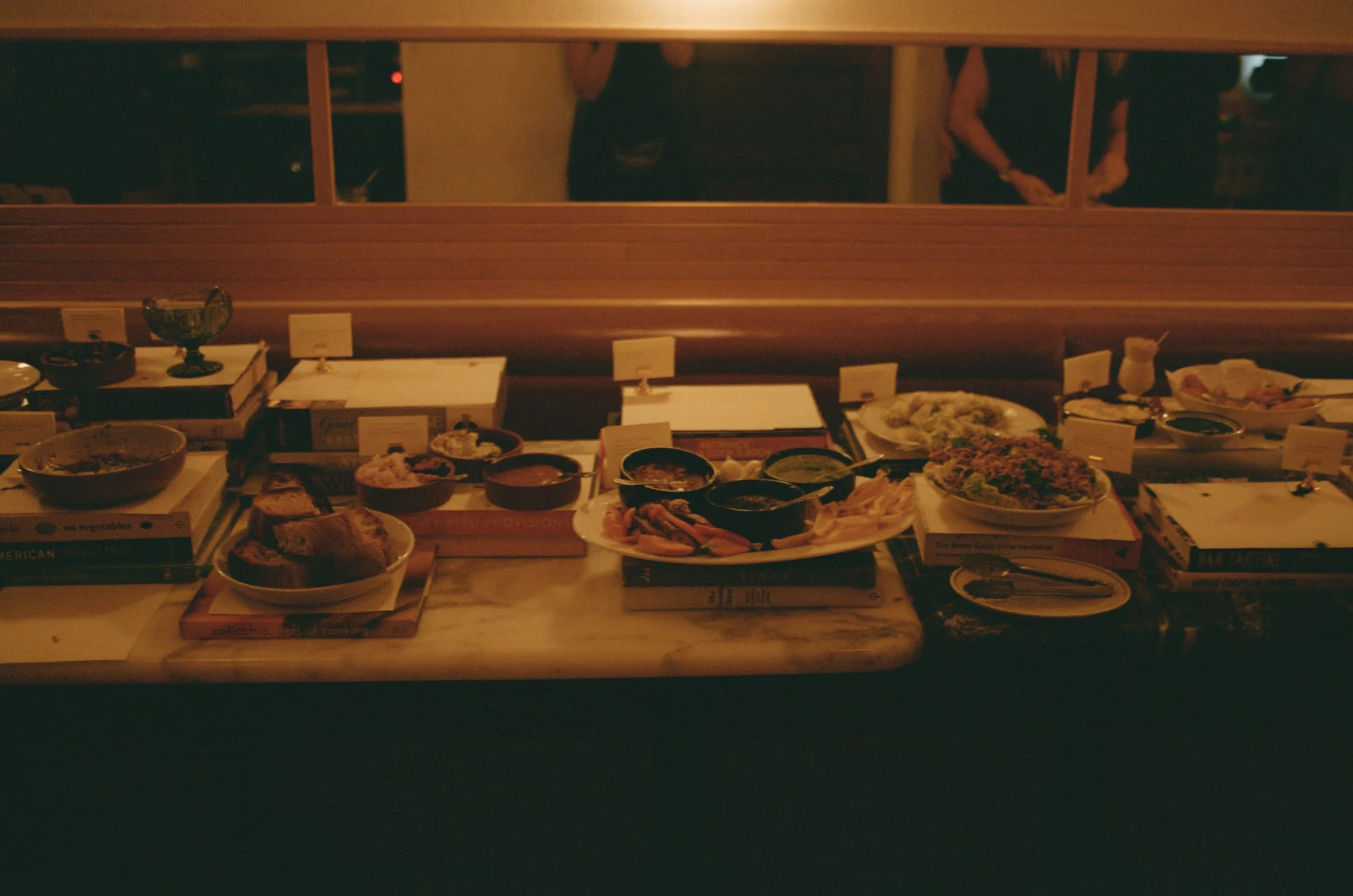 Various dishes and books on a table in a dimly lit restaurant, with a mirror reflecting people in the background.
