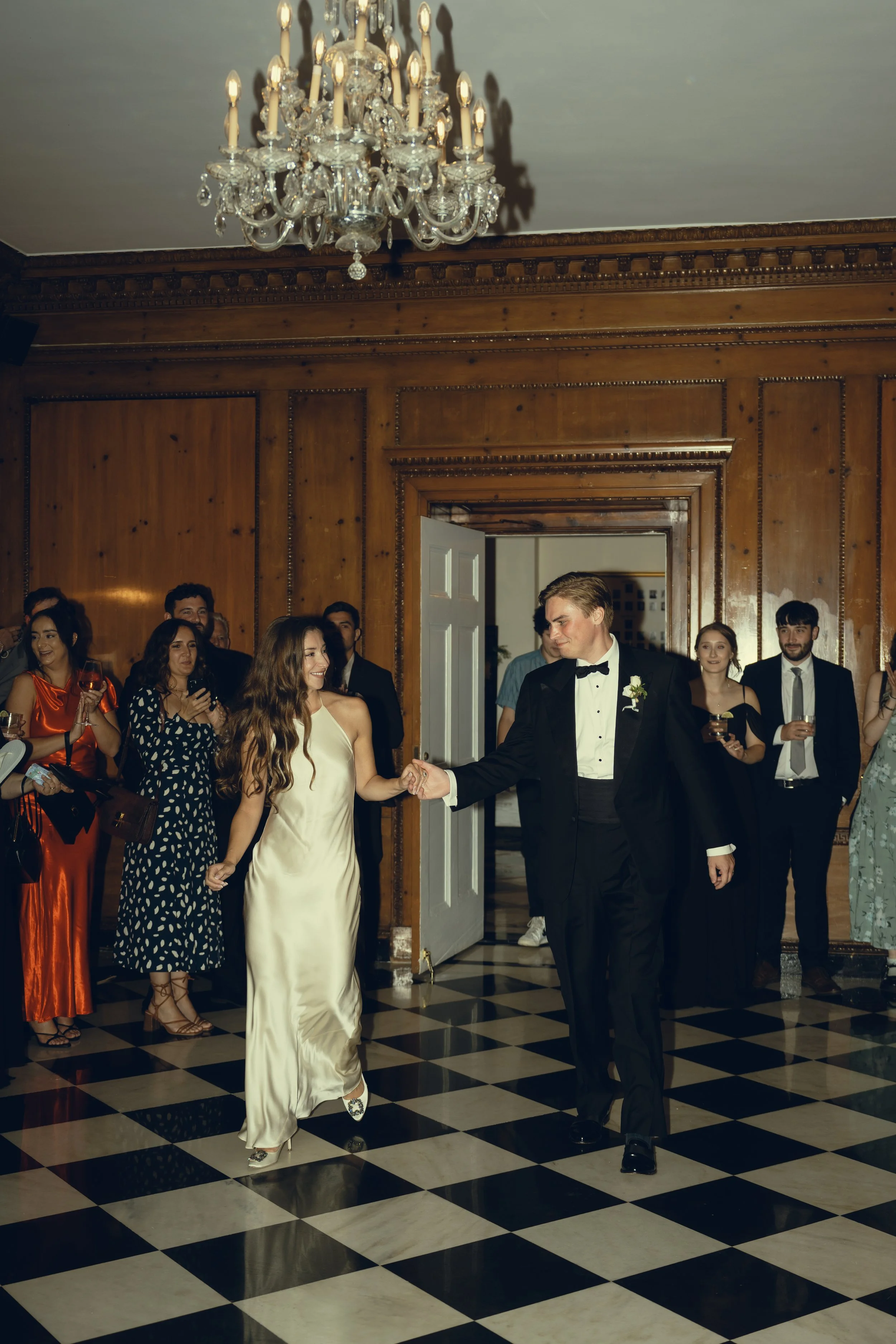 A wedding reception with a couple dancing in the center, surrounded by guests in formal attire, in an elegant room with wood-paneled walls and a chandelier.