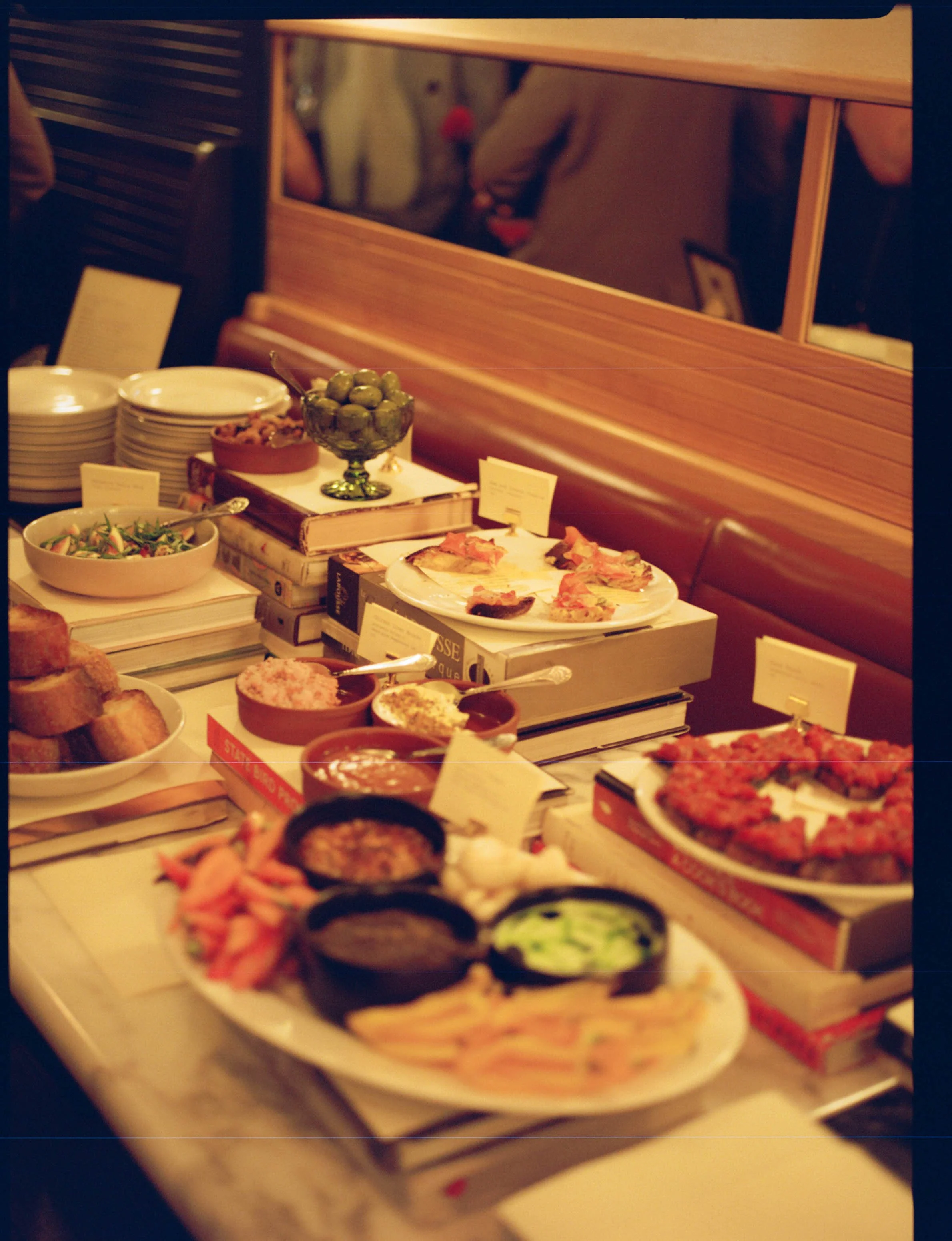 A table set with various dishes including sushi, dips, salads, and appetizers, with books stacked underneath, in a warm, cozy restaurant setting.