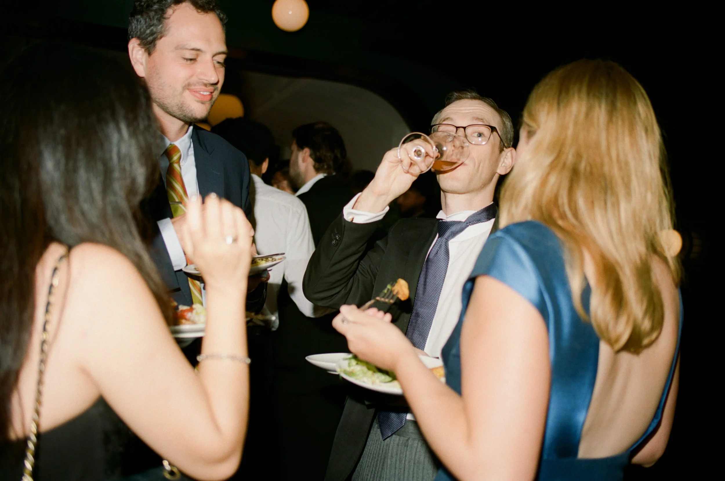 People socializing at a formal event, with a man in a suit drinking from a glass, and others holding plates of food, engaged in conversation.