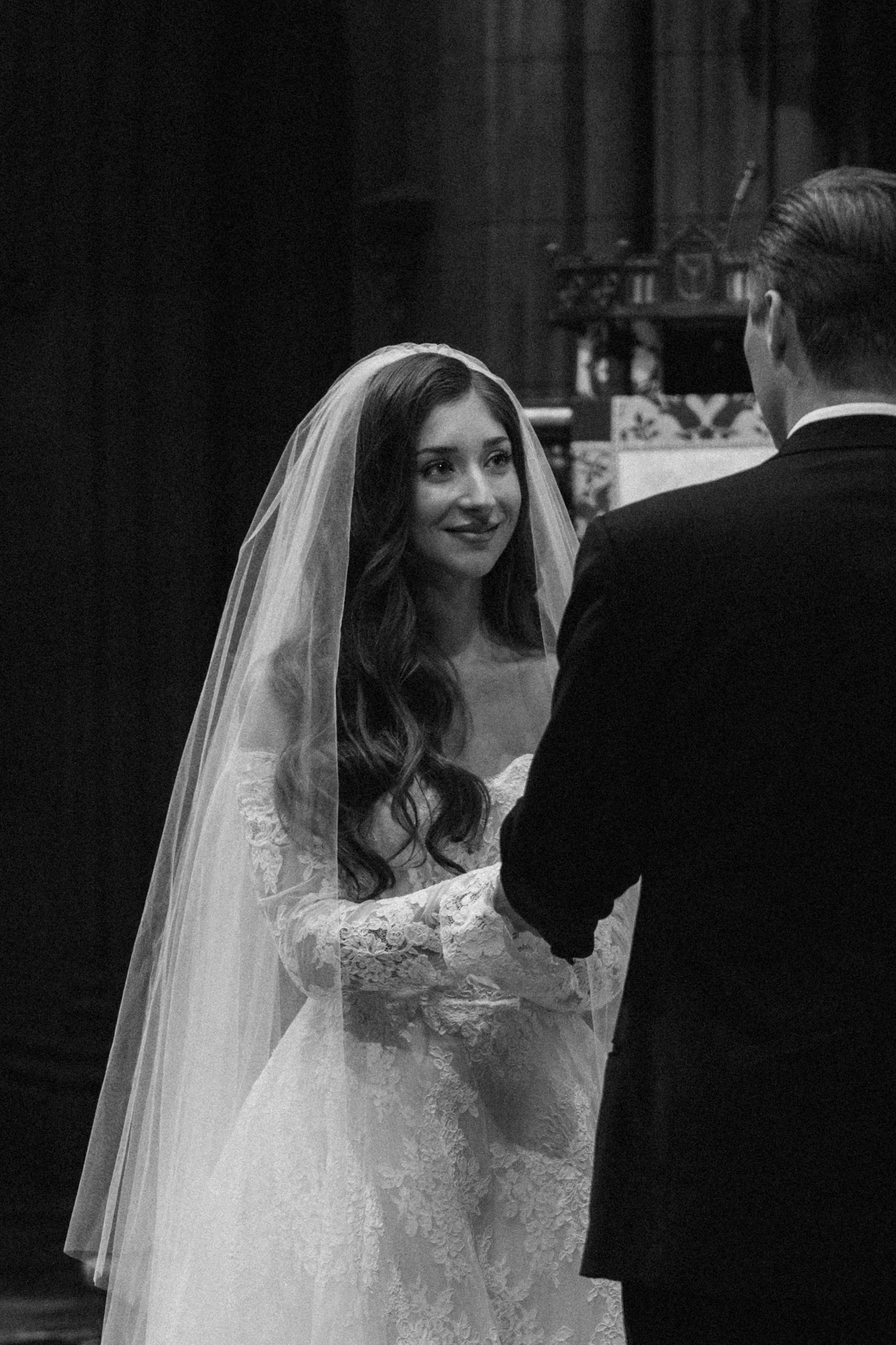 A bride and groom exchange vows at their wedding ceremony, with the bride smiling and wearing a lace wedding dress and veil, while the groom in a suit faces her.