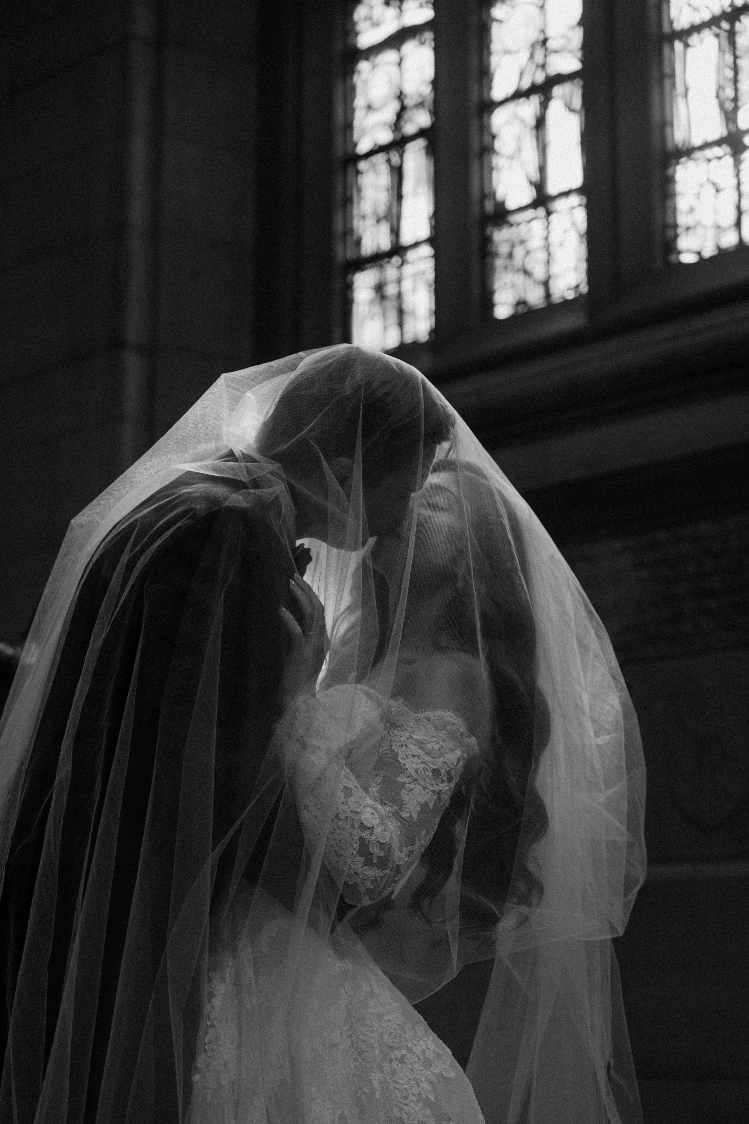 A black and white photo of a bride and groom kissing inside a church, with the bride wearing a lace wedding dress and veil, and the groom in formal attire, illuminated by a large window in the background.