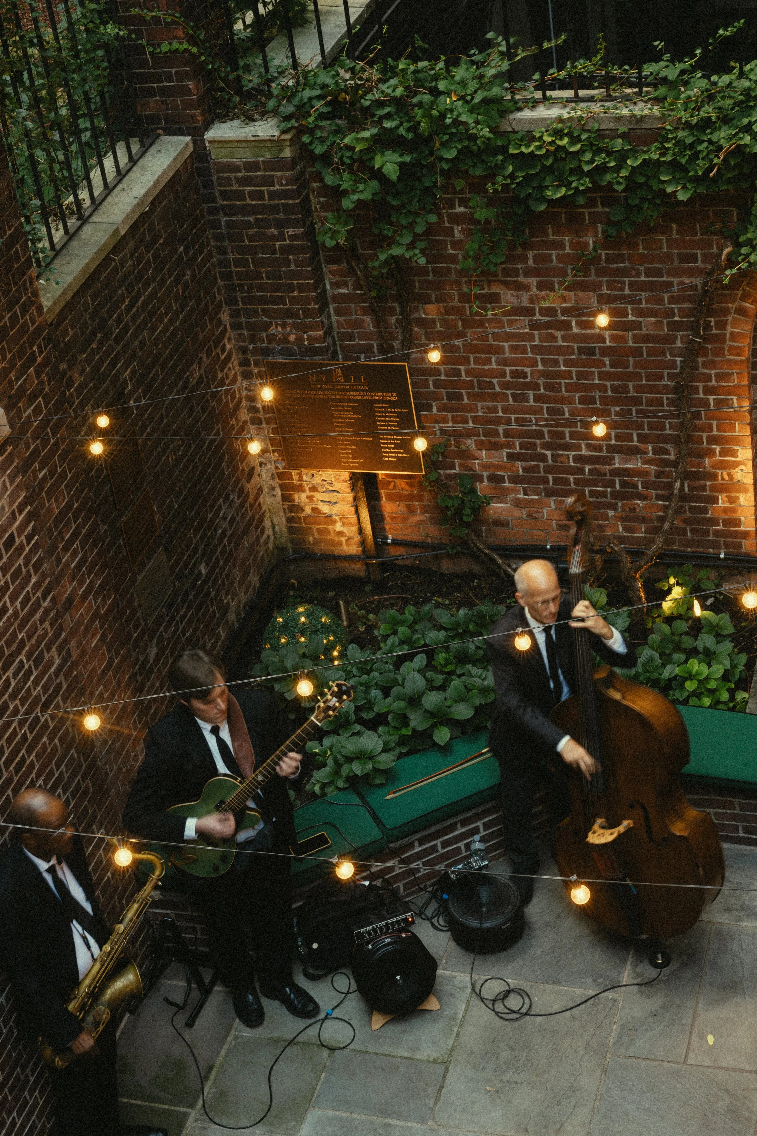 Jazz band performing outdoors on a small stage with string lights above, featuring a guitarist, a double bass player, and a saxophonist against a brick wall with greenery.