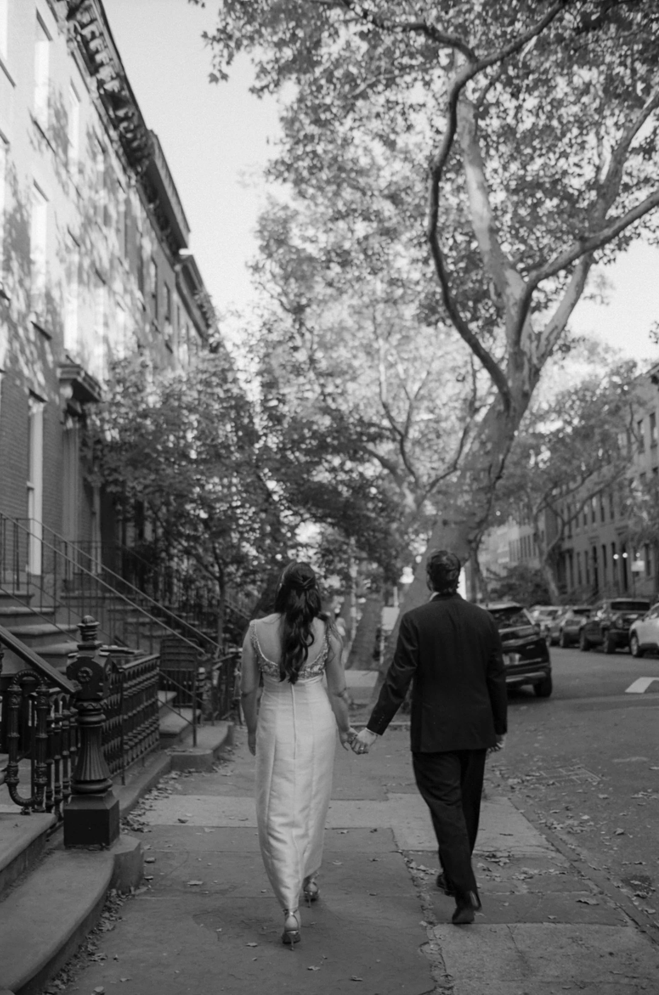 A couple walking hand in hand down a city sidewalk, with trees and residential buildings on either side.