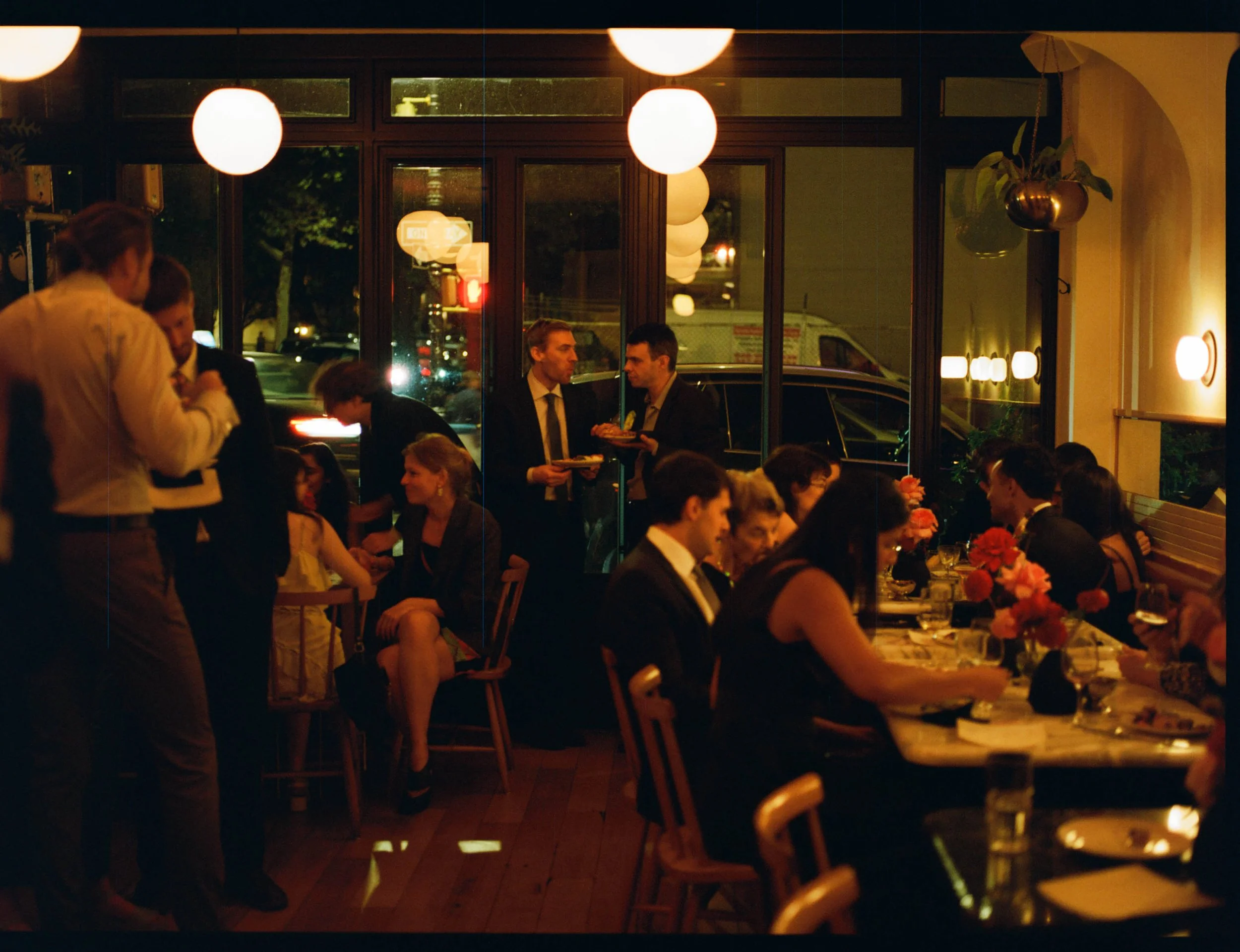 People dining and socializing at a restaurant during evening, with window view of parked cars and street outside, warm lighting, and floral centerpieces on tables.