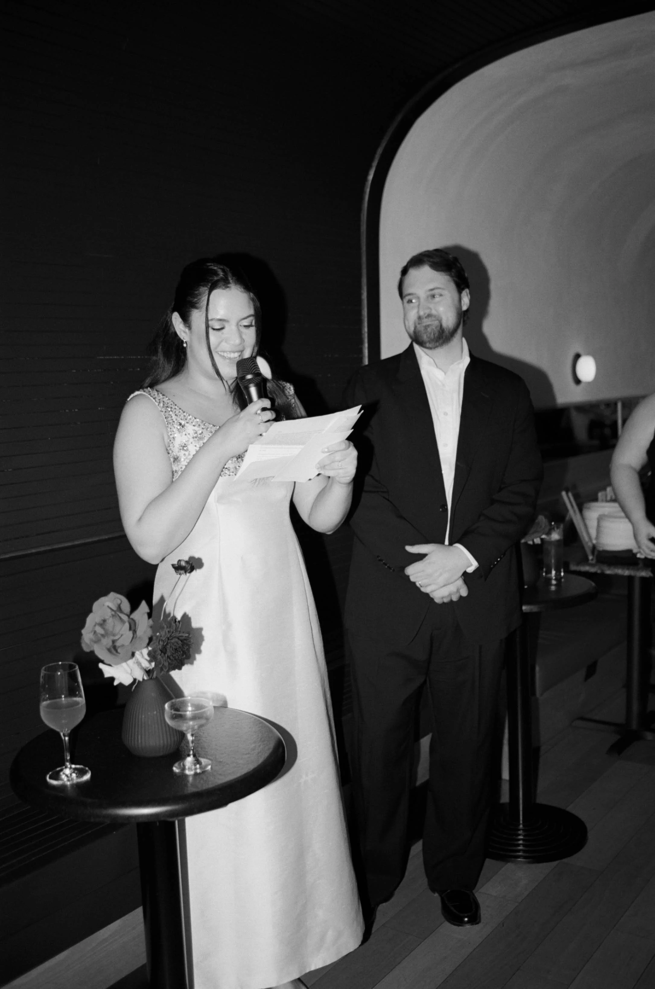 A woman in a white dress speaking into a microphone with a man in a suit standing beside her at a formal event.