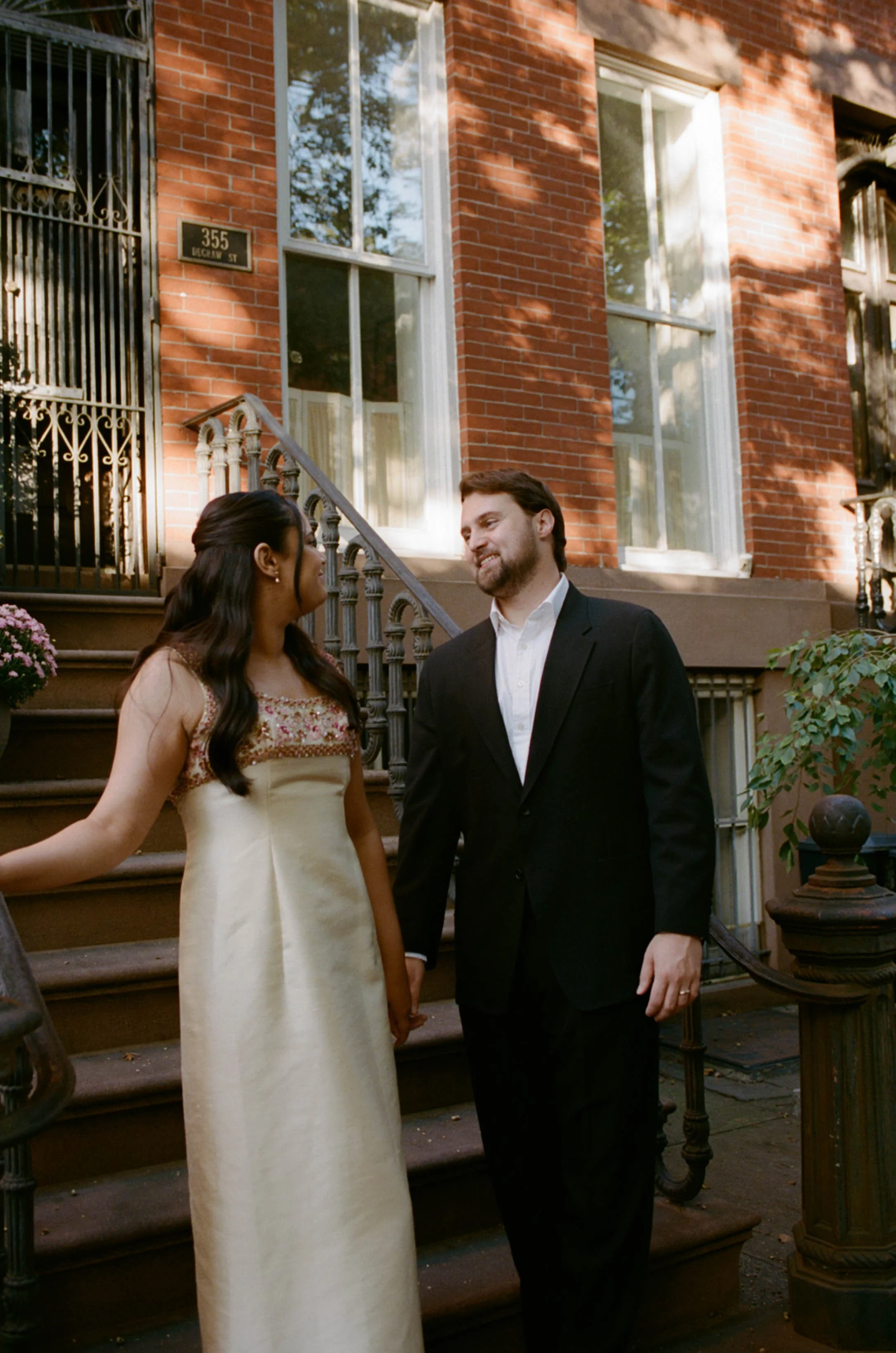 A woman in a cream-colored dress and a man in a black suit holding hands and smiling at each other on the steps outside a brick building.
