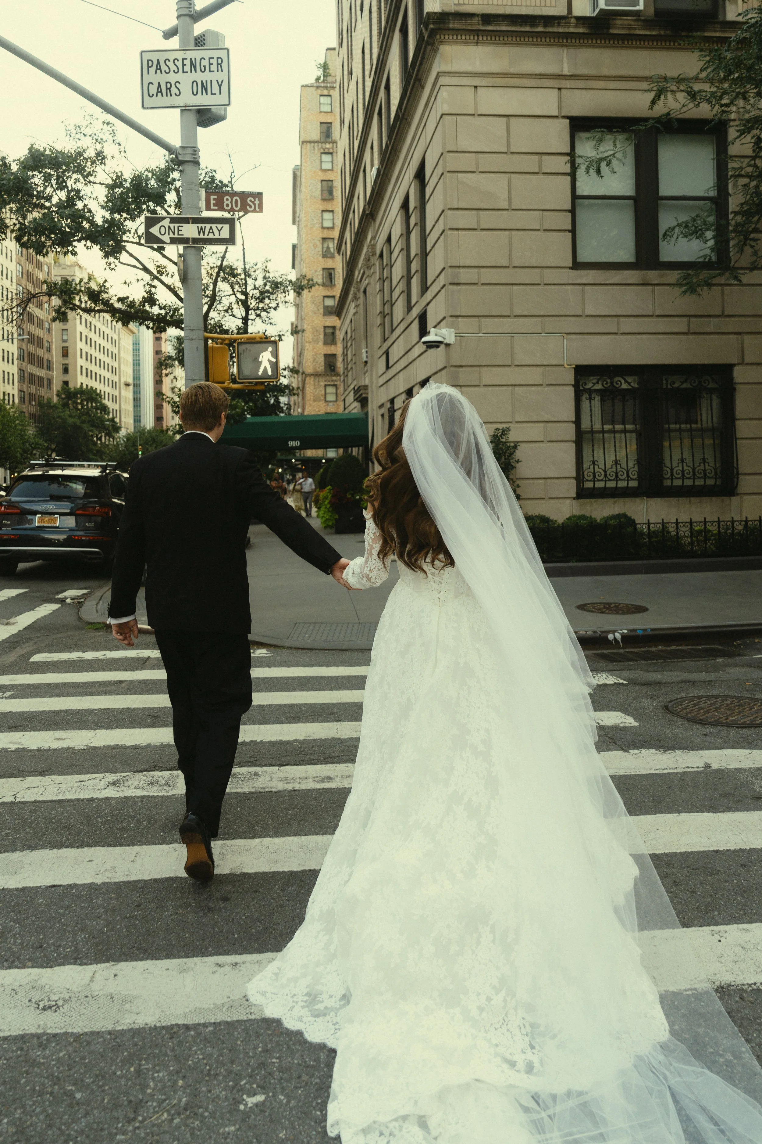 A bride and groom holding hands and walking across a city crosswalk near buildings, cars, and street signs, including a pedestrian crossing signal and a sign indicating one-way street.