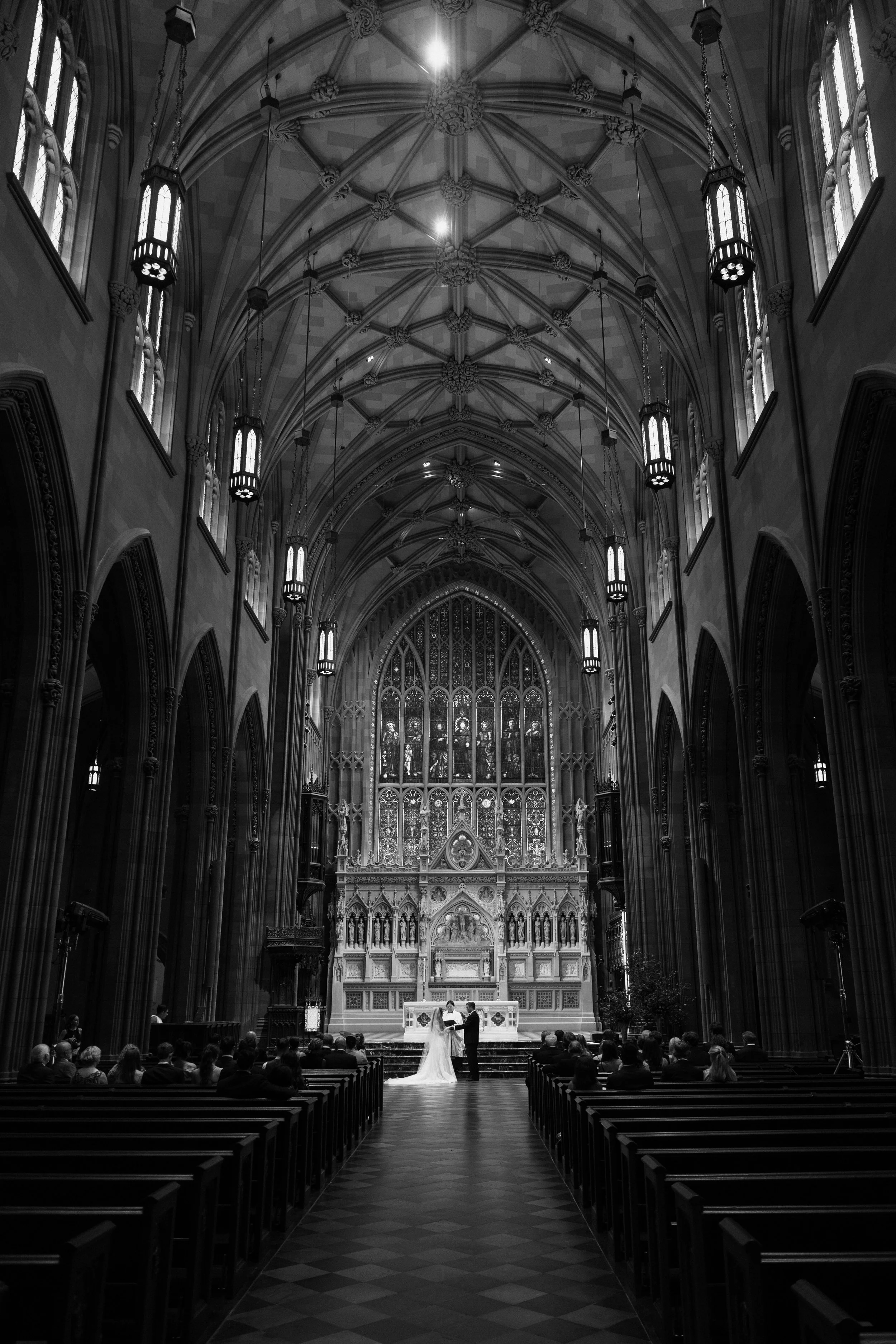 Wedding ceremony taking place inside a Gothic-style cathedral with high vaulted ceilings, stained glass windows, and ornate architecture, with a bride and groom at the altar and guests seated in pews.