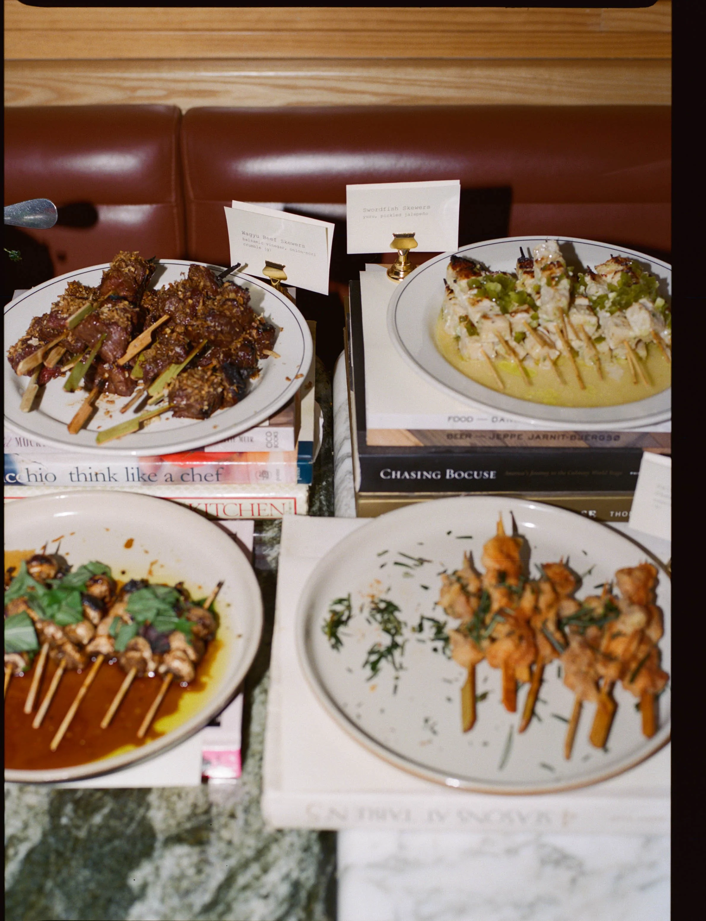 Assorted Asian appetizers on white plates displayed on a table with books in the background. Includes skewers of glazed meat, a dish with slices of fish in a sauce, grilled skewers with herbs, and another dish with fried pieces garnished with herbs.