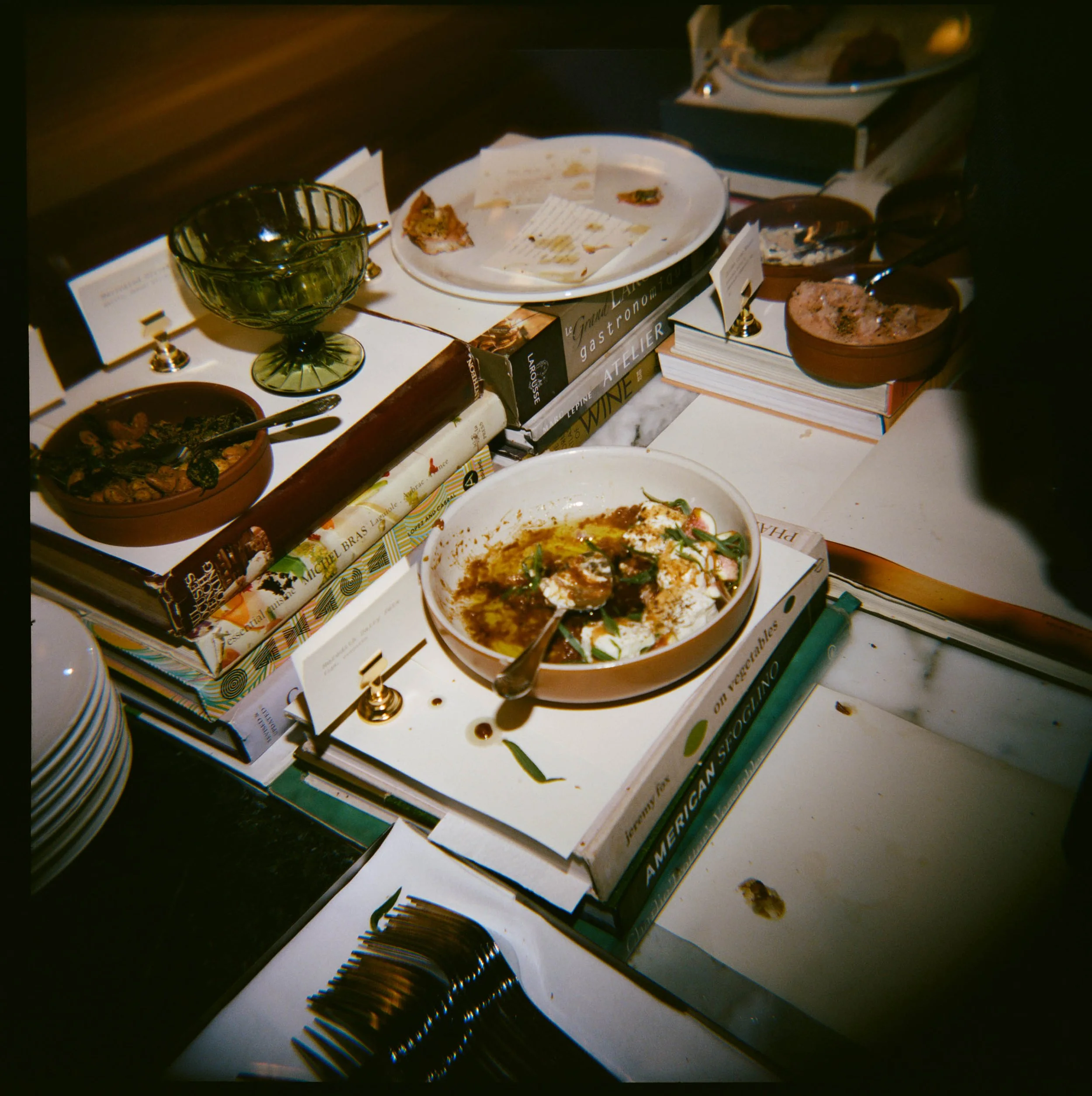 A cluttered table with dishes, books, and utensils, including a bowl of partially eaten food, a green glass bowl, and stacks of books.