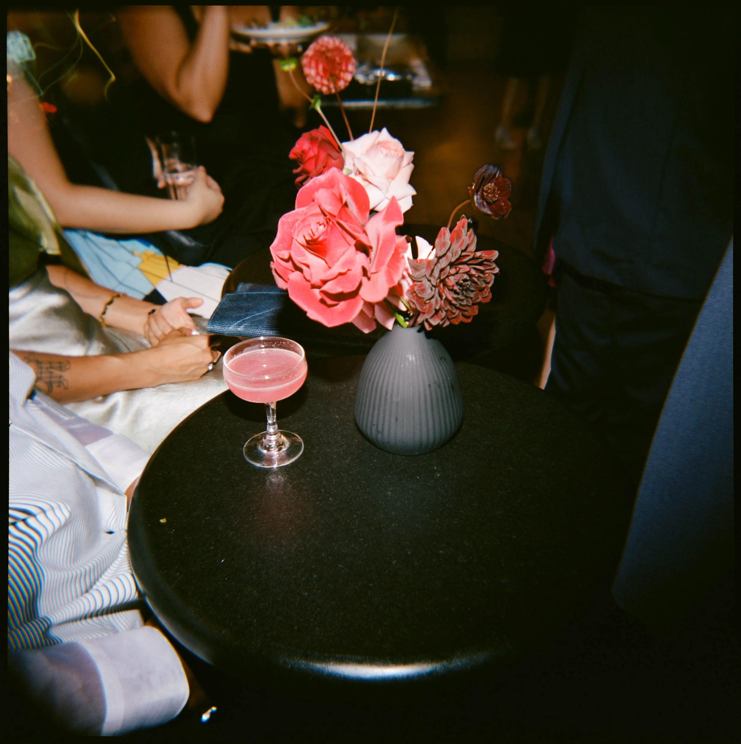A black table with a matte gray vase holding pink, red, and brown artificial flowers, accompanied by a pink cocktail in a coupe glass, with people sitting around in a dimly lit setting.