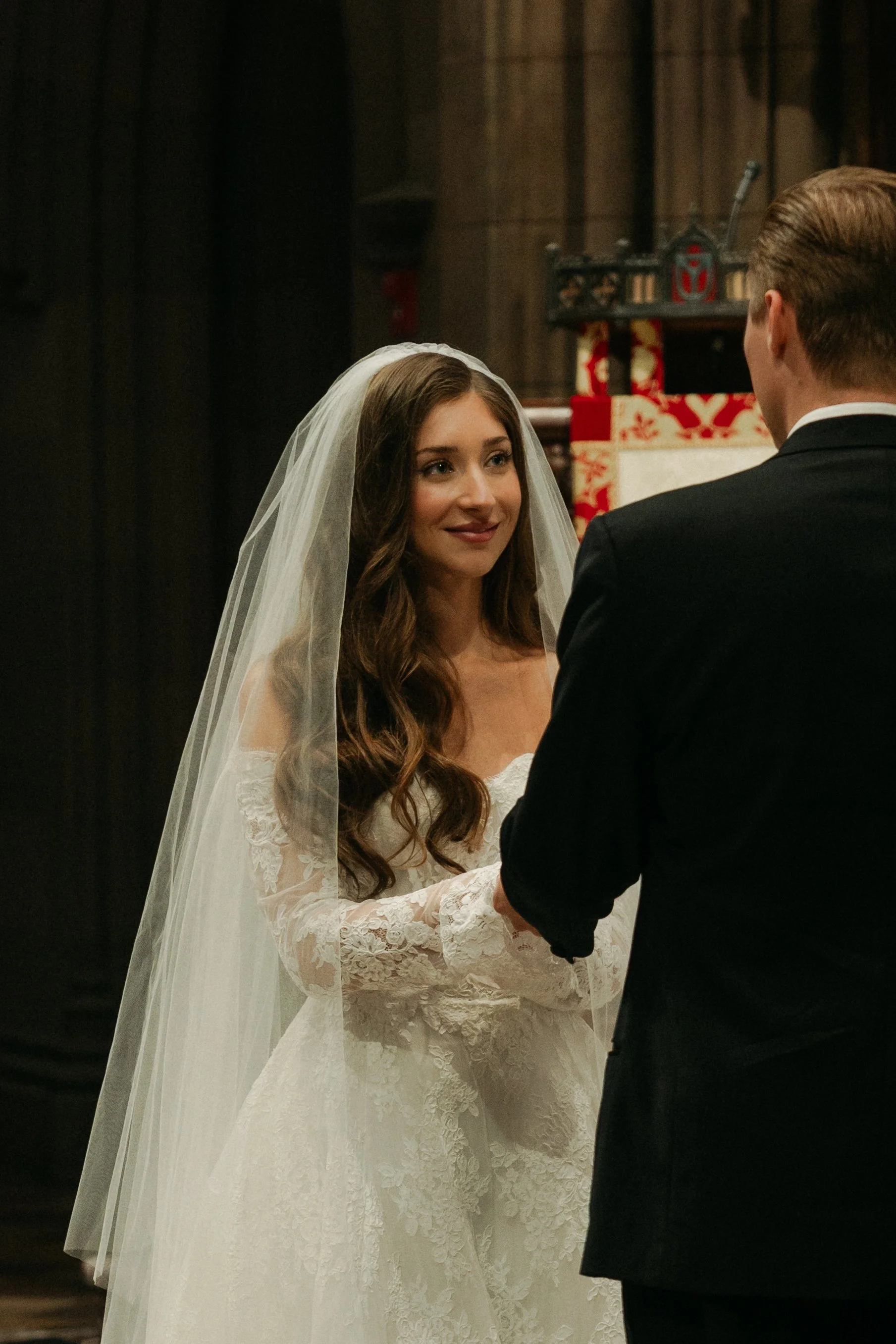 A bride and groom holding hands during their wedding ceremony, with the bride looking at him lovingly inside a church.