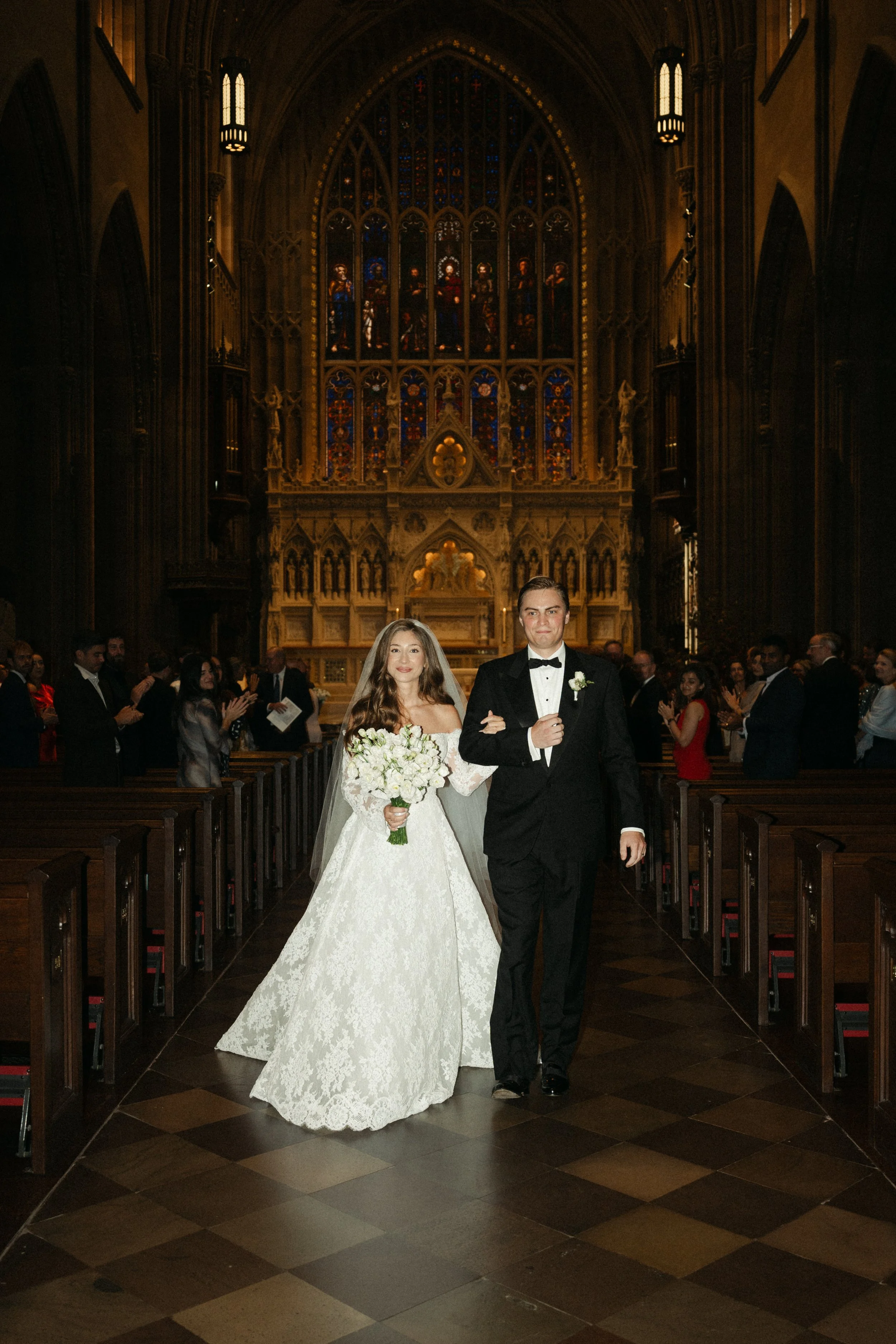 A newlywed couple walks down the aisle inside a church, with the bride holding a bouquet of white flowers and the groom in a black tuxedo with a bow tie, surrounded by wedding guests.